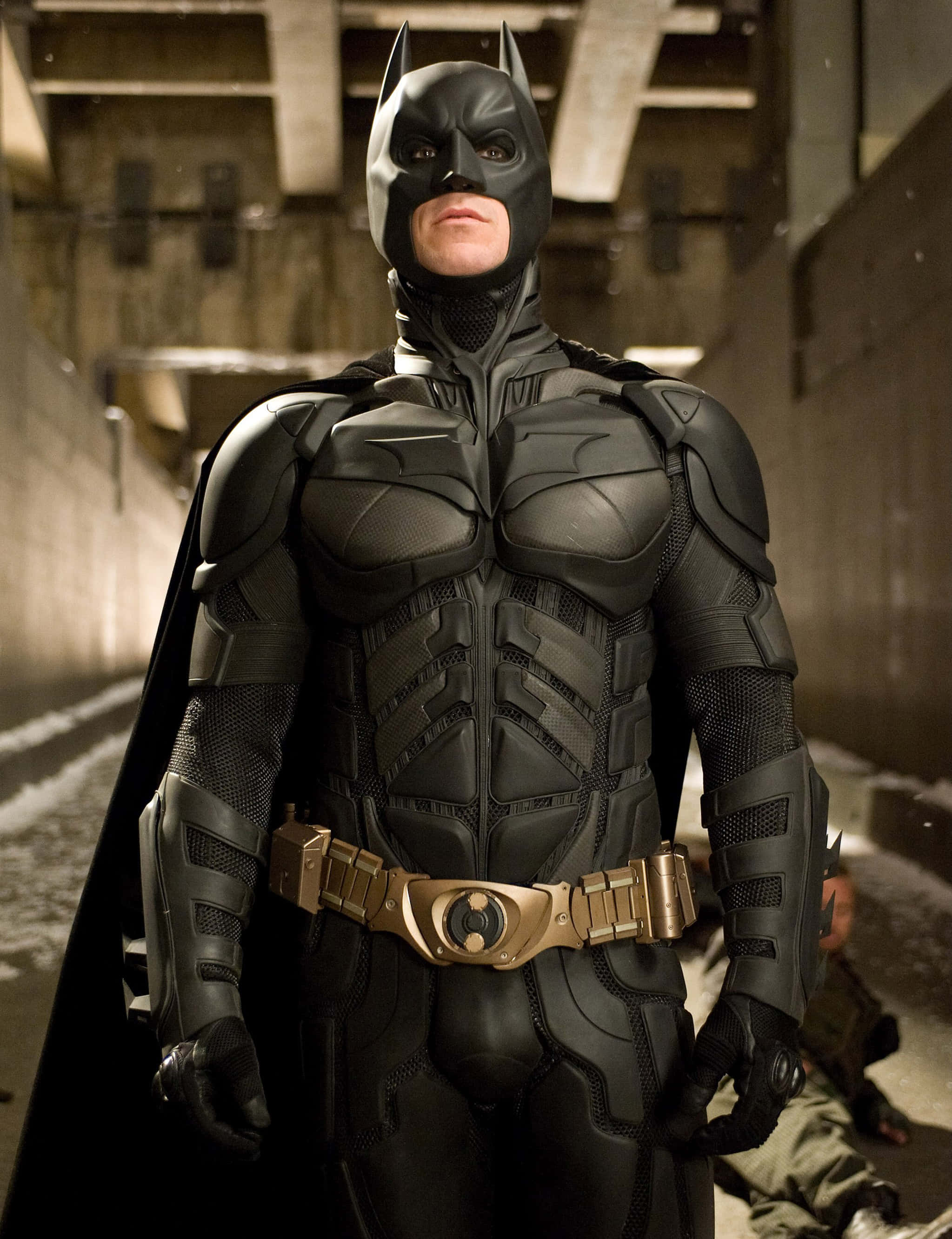 Bat-suit 桌布 壁紙 — 蝙蝠俠身穿裝甲服，在昏暗的混凝土拱門下巍然站立，腳下有一個倒下的身影。