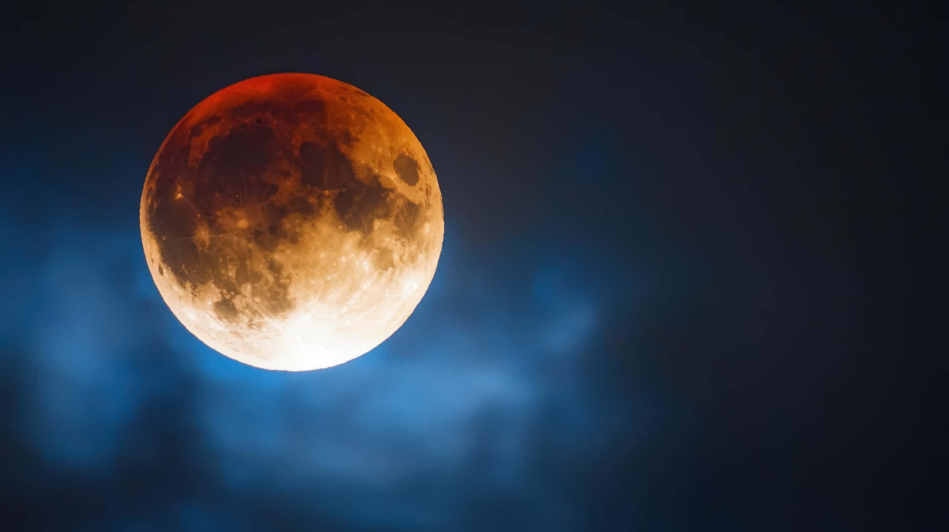 Blood Moon Lunar Eclipse in Night Sky with Dramatic Clouds 壁紙 — 血月發出琥珀色和橙色的光芒，映襯著深藍色的夜空和縷縷雲彩。