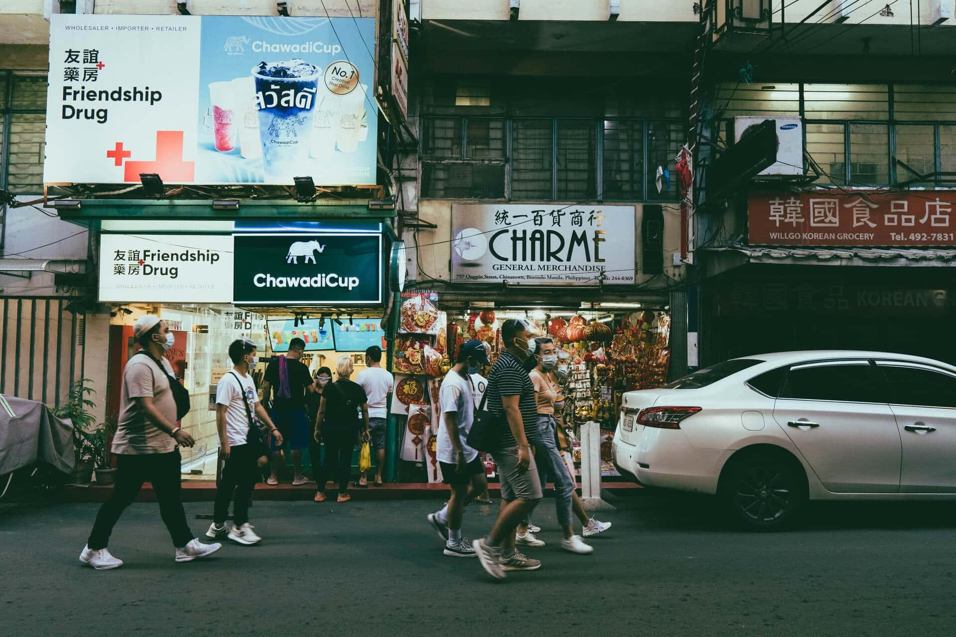 Street 桌布 壁紙 — 行人在溫暖的晚燈下穿過一條繁華的城市街道，兩旁是燈火通明的商店櫥窗和多語言的招牌。