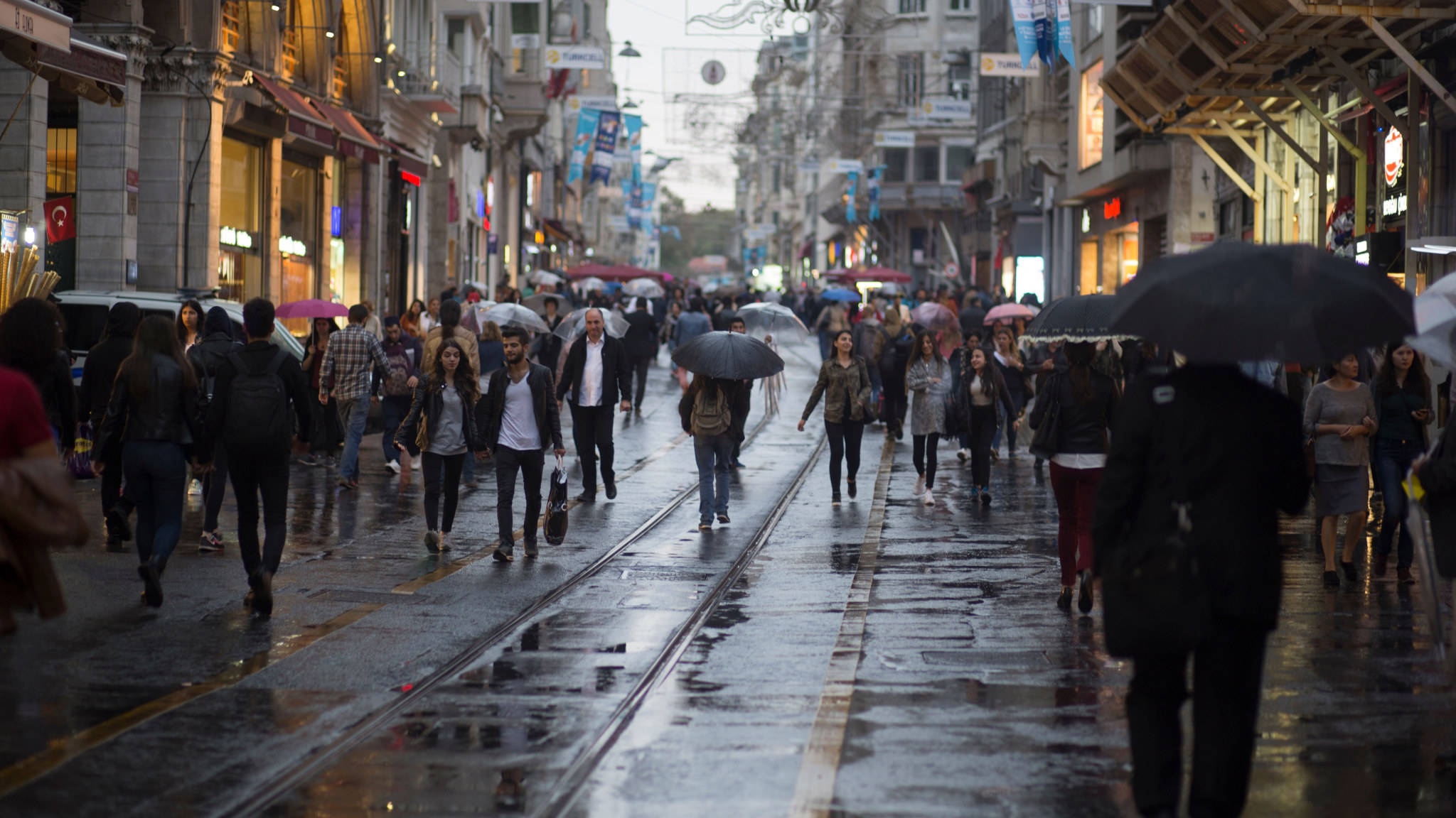 人物 桌布 壁紙 — 行人撐著雨傘，在陰天之下，沿著兩旁商店和電車軌道的濕滑城市街道前行。