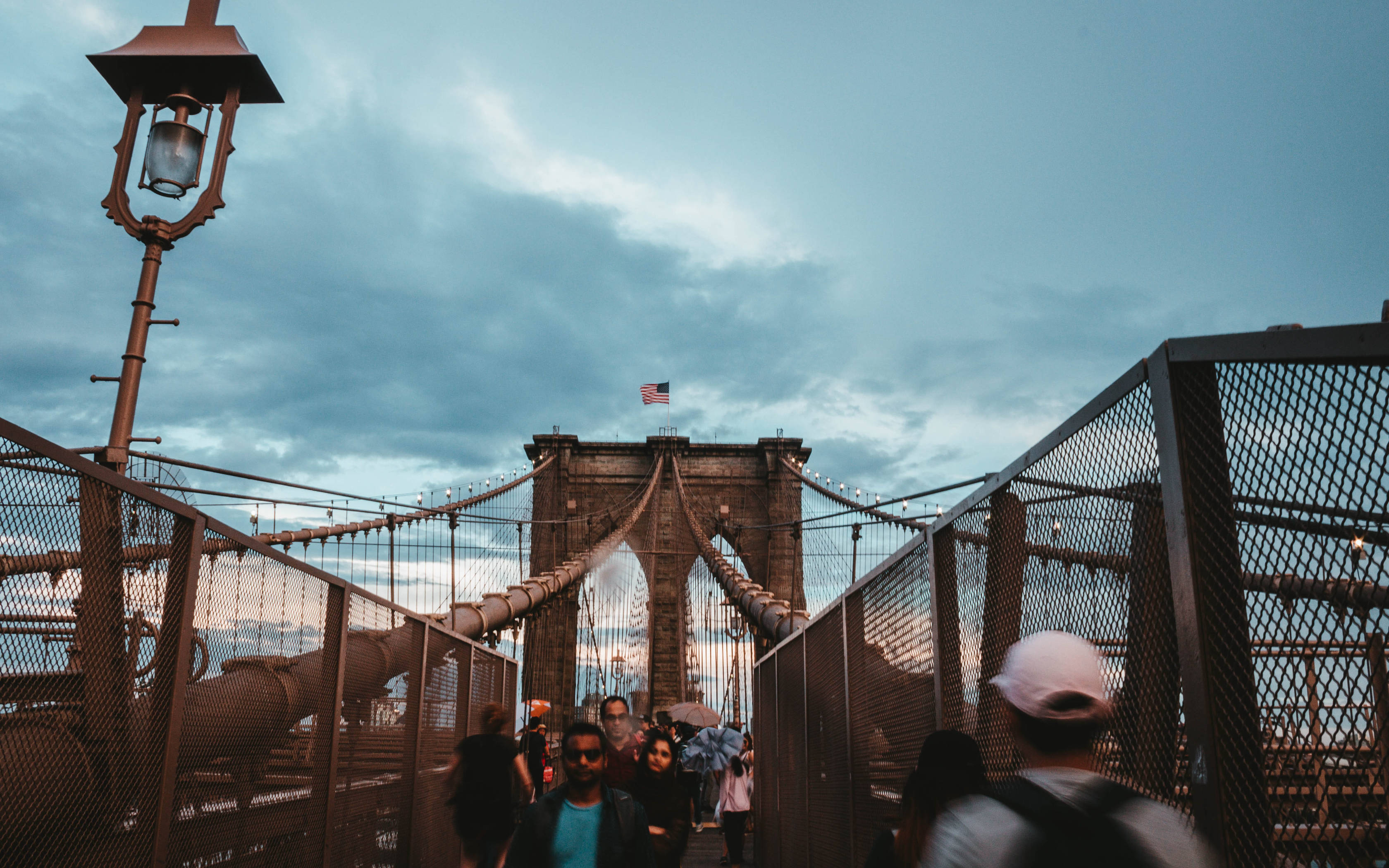 New York 桌布 壁紙 — 行人漫步在標誌性的布魯克林大橋上，暮色蒼茫，美國國旗在其石塔頂端飄揚。
