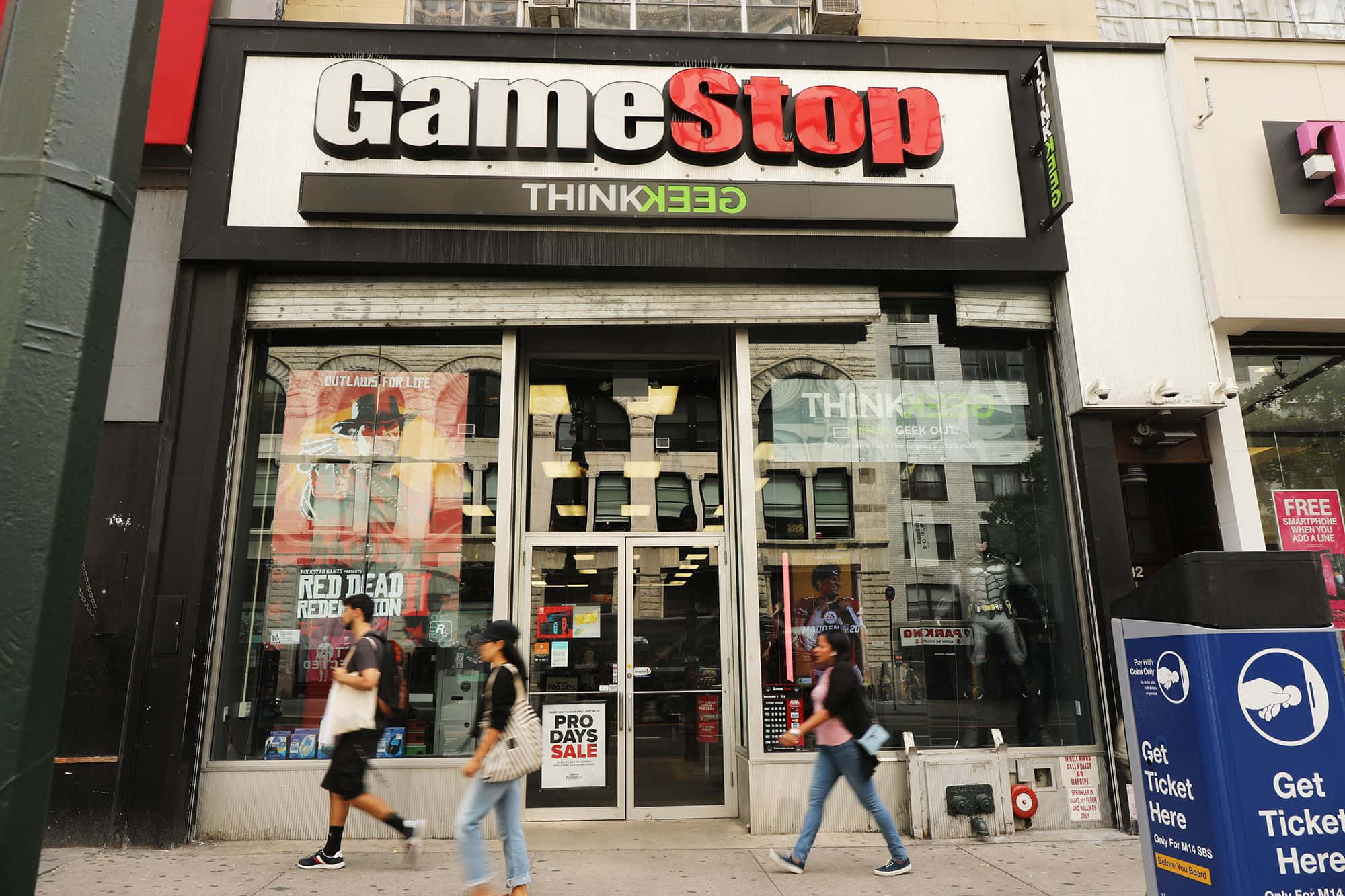 GameStop 桌布 壁紙 — 行人經過一間GameStop店鋪，玻璃窗內可見醒目的招牌和遊戲海報。