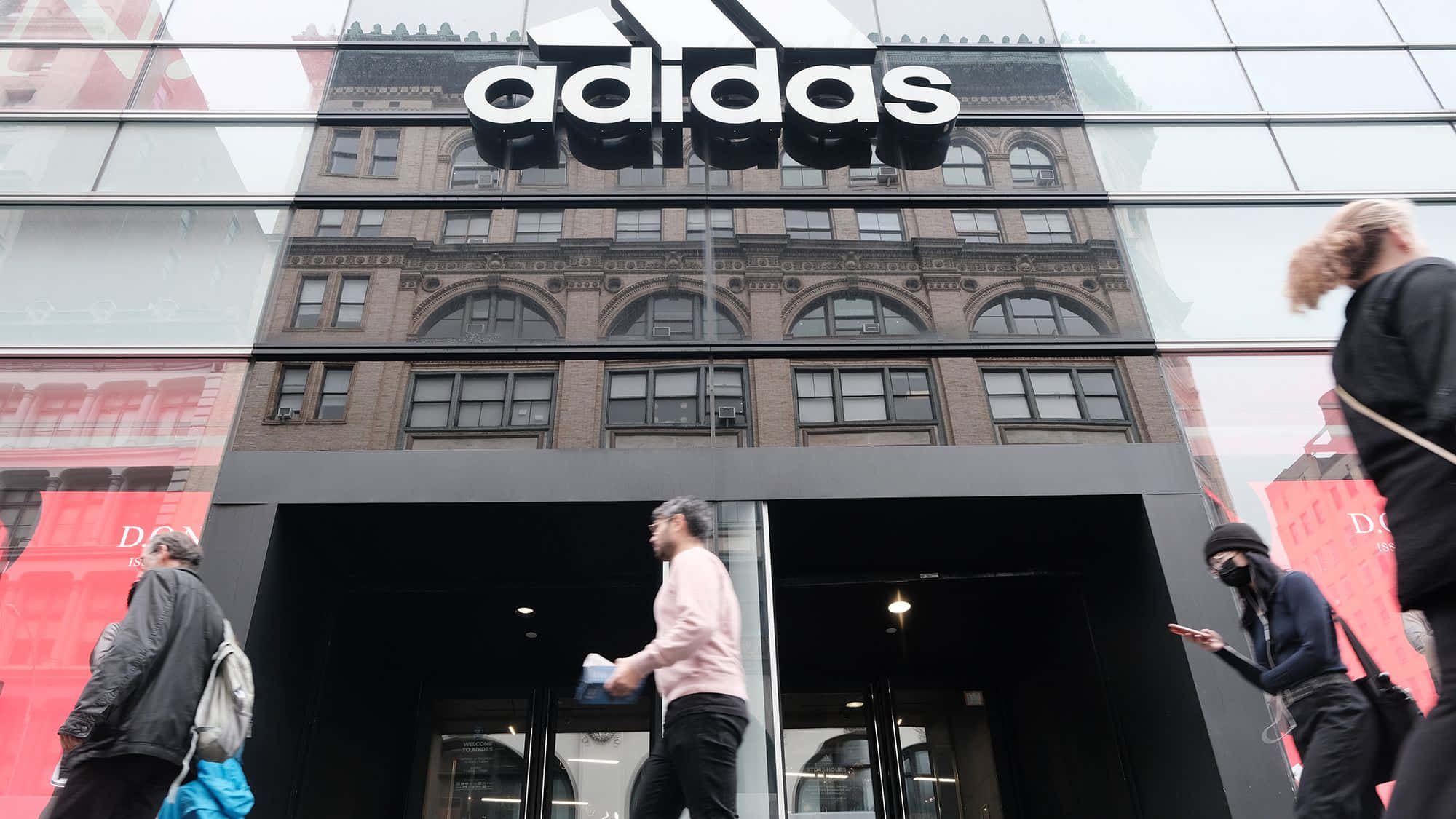 愛迪達 桌布 壁紙 — 行人走過一間現代化的adidas商店，玻璃外牆反射出背後歷史悠久的磚建築。