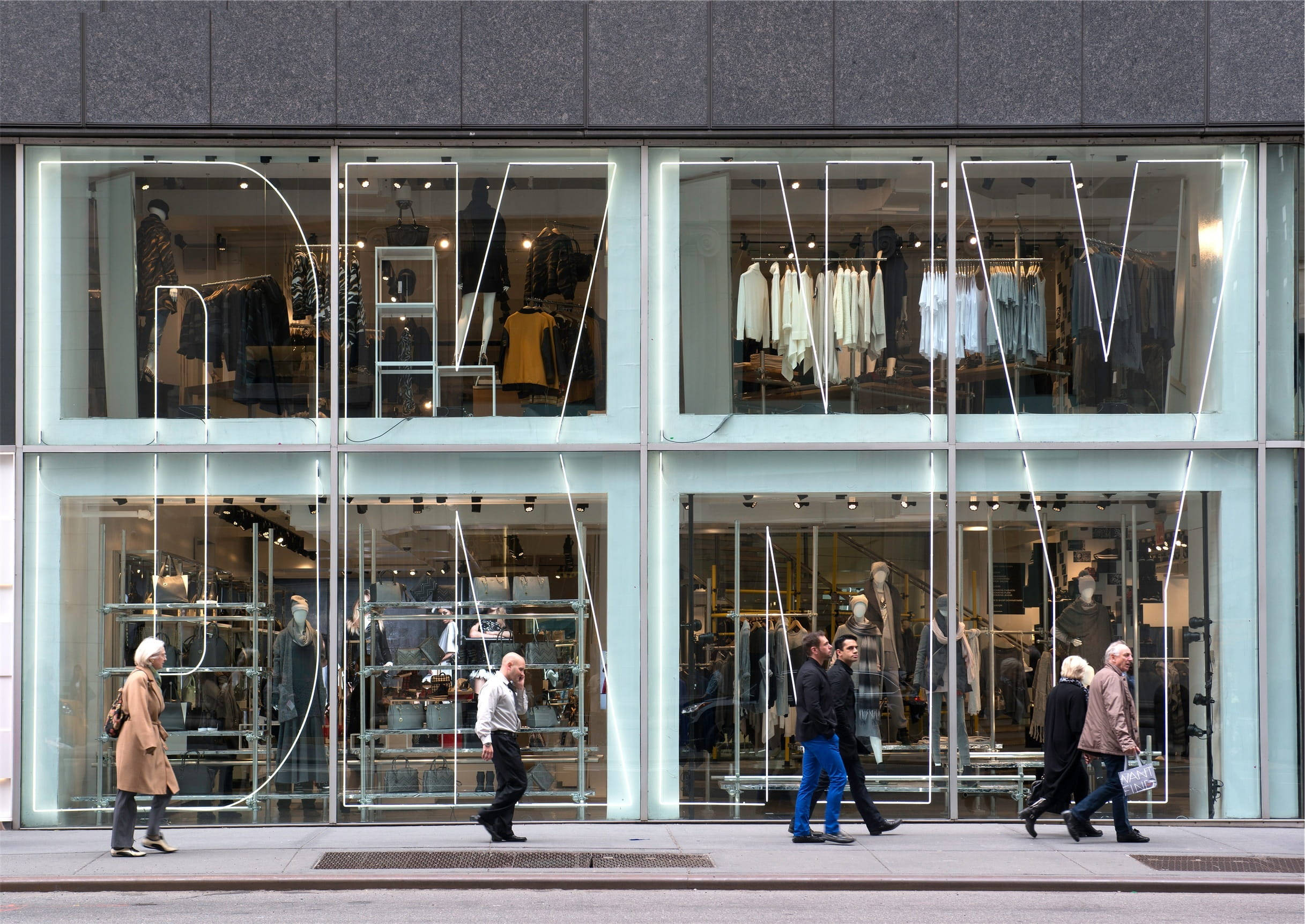 品牌 桌布 壁紙 — 行人走過一間現代玻璃幕牆服裝店，店內有發光的「DKNY」標誌和可見的服裝架。