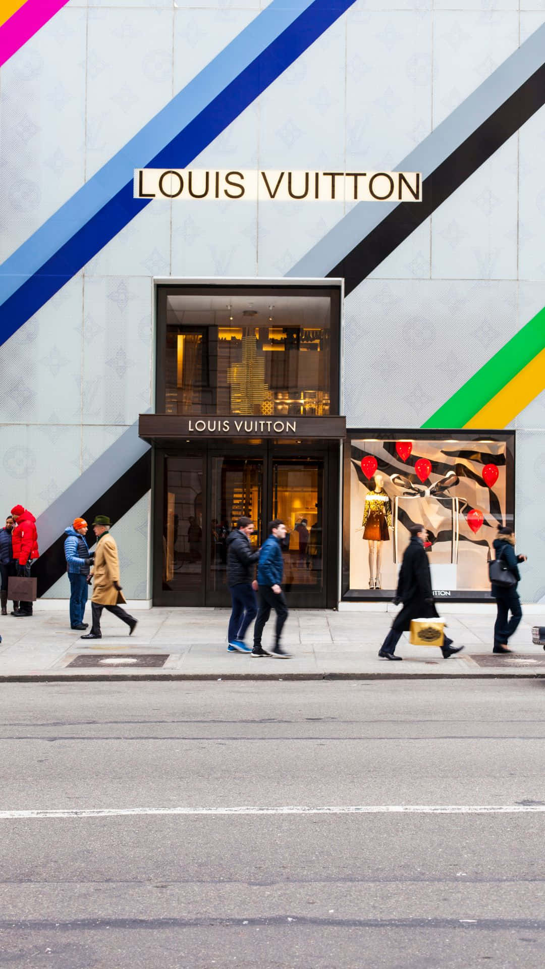 louis vuitton phone 桌布 壁紙 — 行人走過一間路易威登店鋪，店鋪有鮮明的對角線條和發光的櫥窗，位於繁華的街道上。