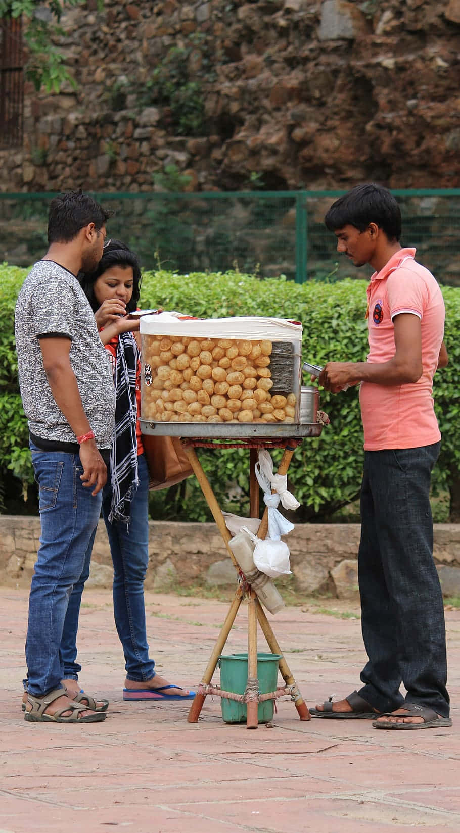 Indian 桌布 壁紙 — 街頭小販在戶外攤位為一對情侶提供金黃色炸小吃，背景有石牆和綠色籬笆。