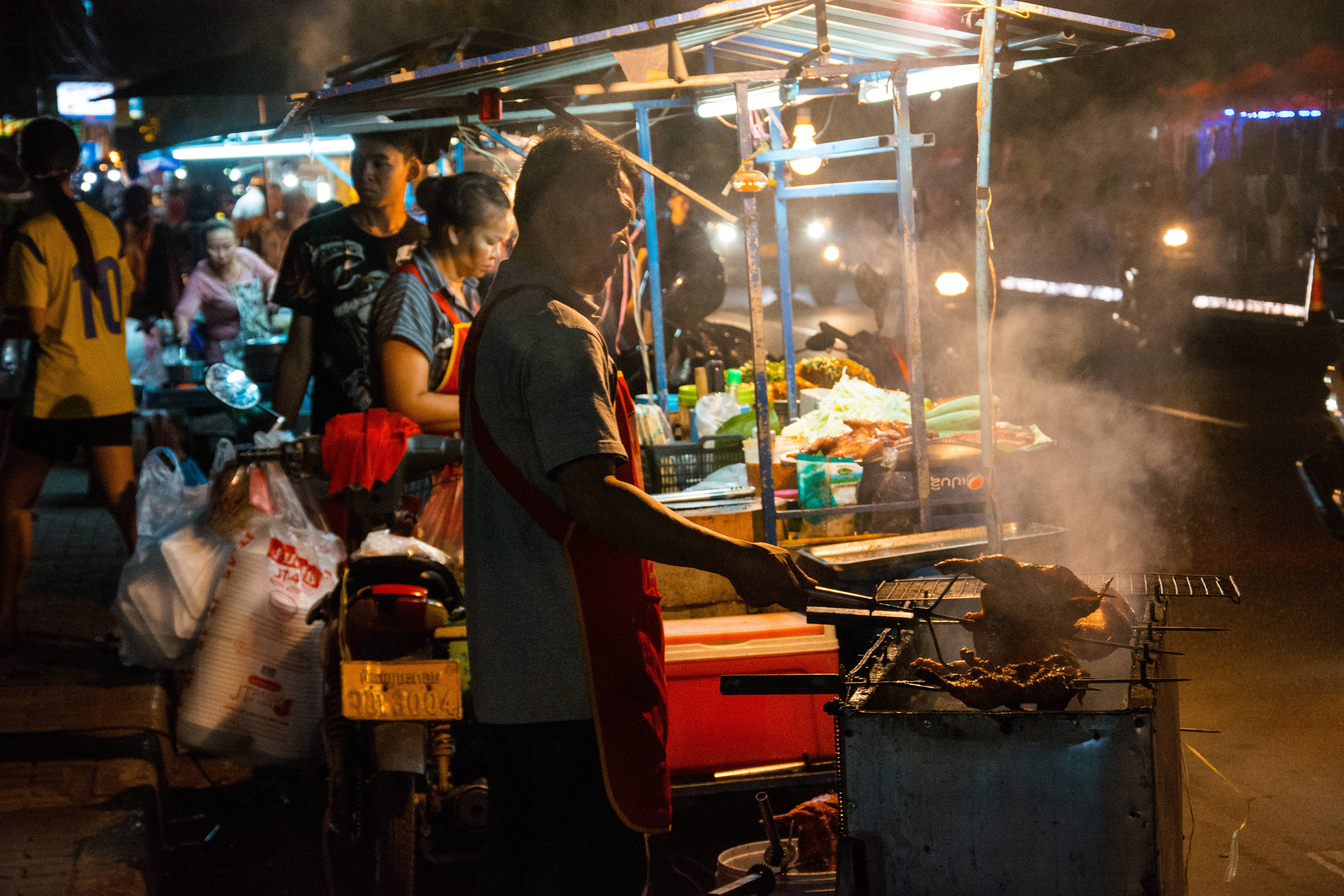 Laos 桌布 壁紙 — 街頭小販在熱鬧的夜市中用炭火烤肉，蒸氣升騰，溫暖的燈光照亮整個場景。