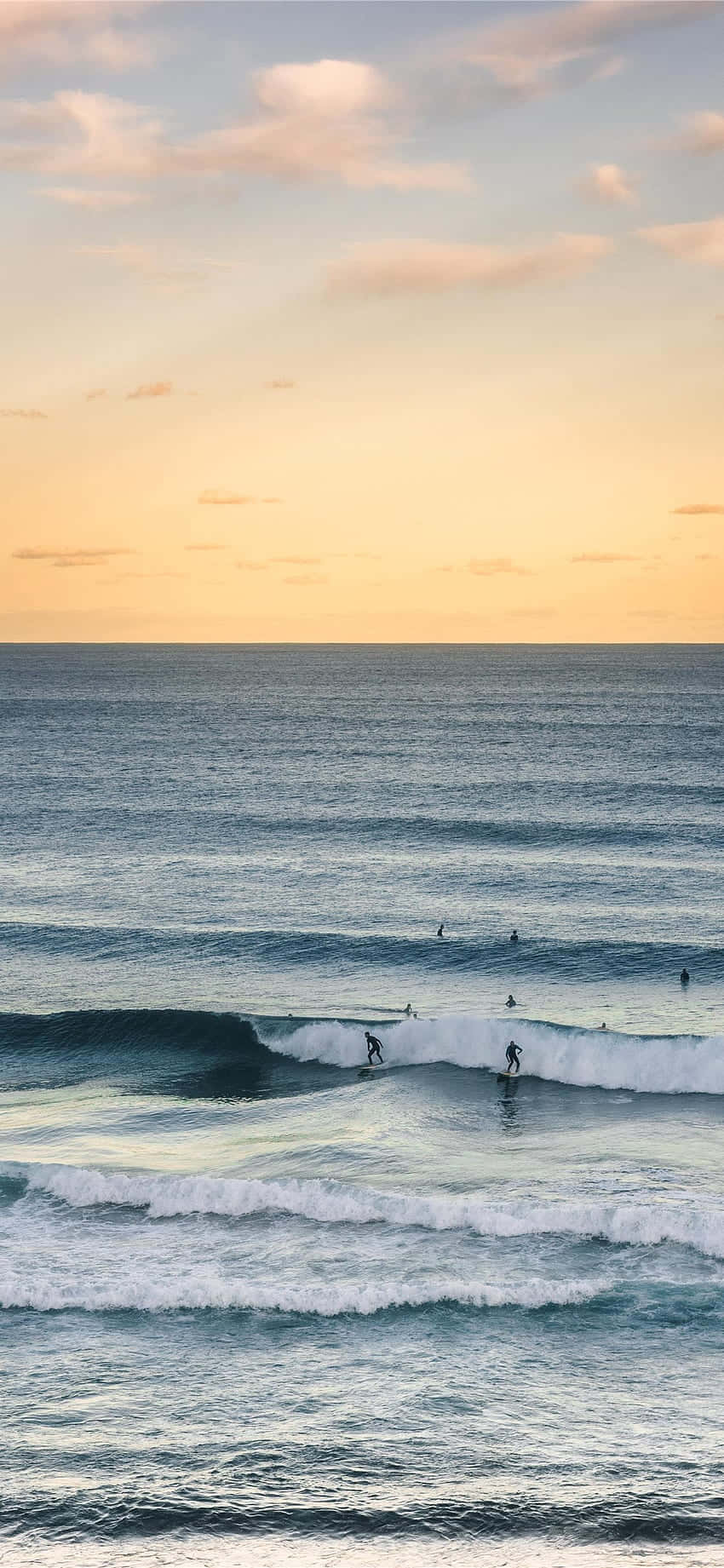 Surfing iPhone 桌布 壁紙 — 衝浪者在柔和的金色日落天空下，乘著散雲，馳騁於海浪之上。