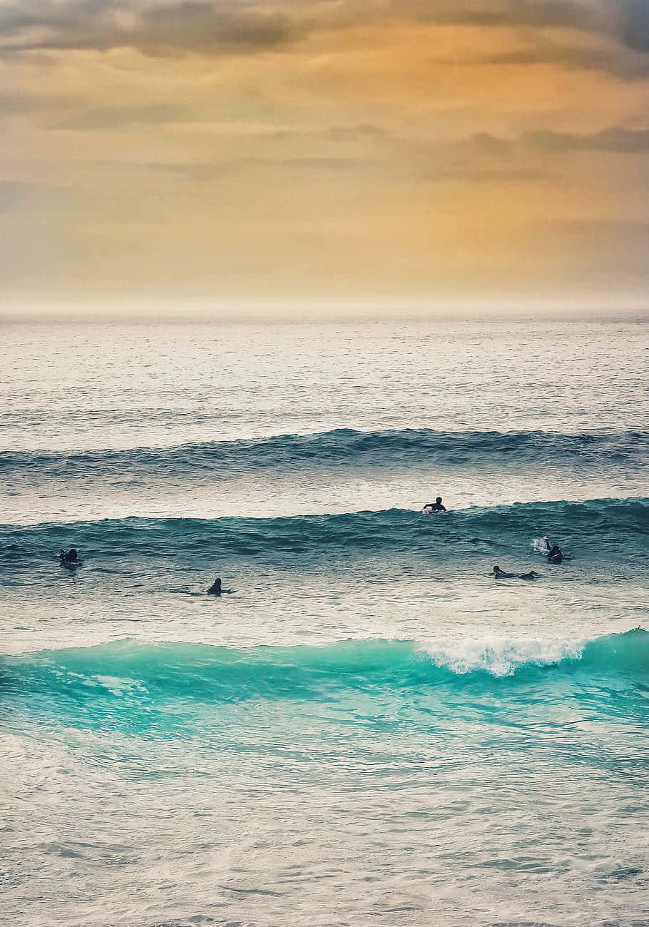 Surfing iPhone 桌布 壁紙 — 衝浪者在柔軟雲朵的金色日落天空下，在碧綠色的海浪上划槳。
