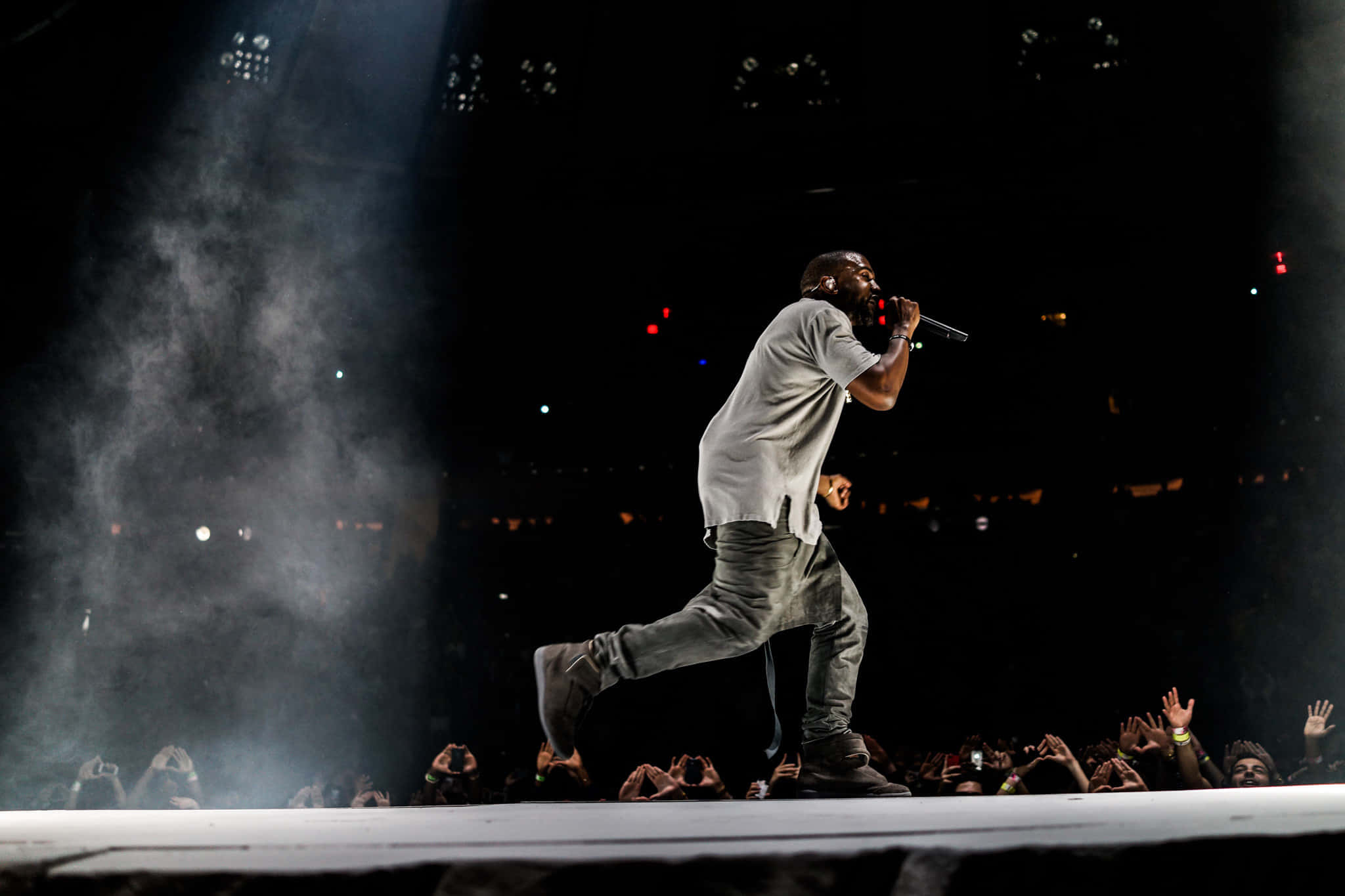 Kanye West Yeezus 桌布 壁紙 — 表演者在明亮的聚光燈下，在煙霧瀰漫的舞台上動態地大步走過，下方熱情的觀眾伸手向上。