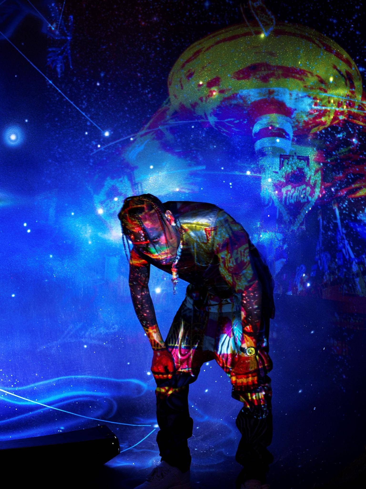 Travis Scott Astroworld 桌布 壁紙 — 表演者在星光閃爍的藍色舞台上，在充滿活力的宇宙投影和發光霓虹圖案下向前彎腰。