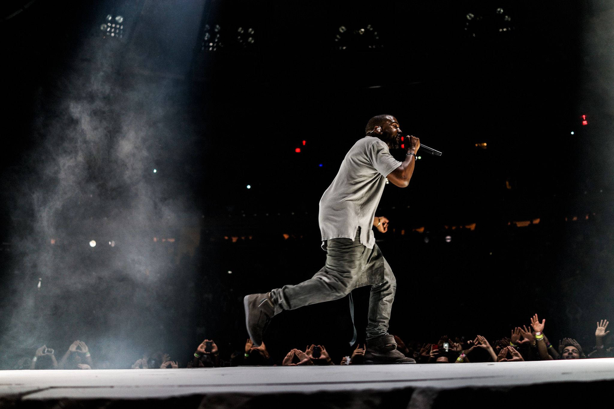 Kanye West 桌布 壁紙 — 表演者在煙霧瀰漫的舞台上，在明亮的聚光燈下動態地大步走，下方有投入的觀眾。