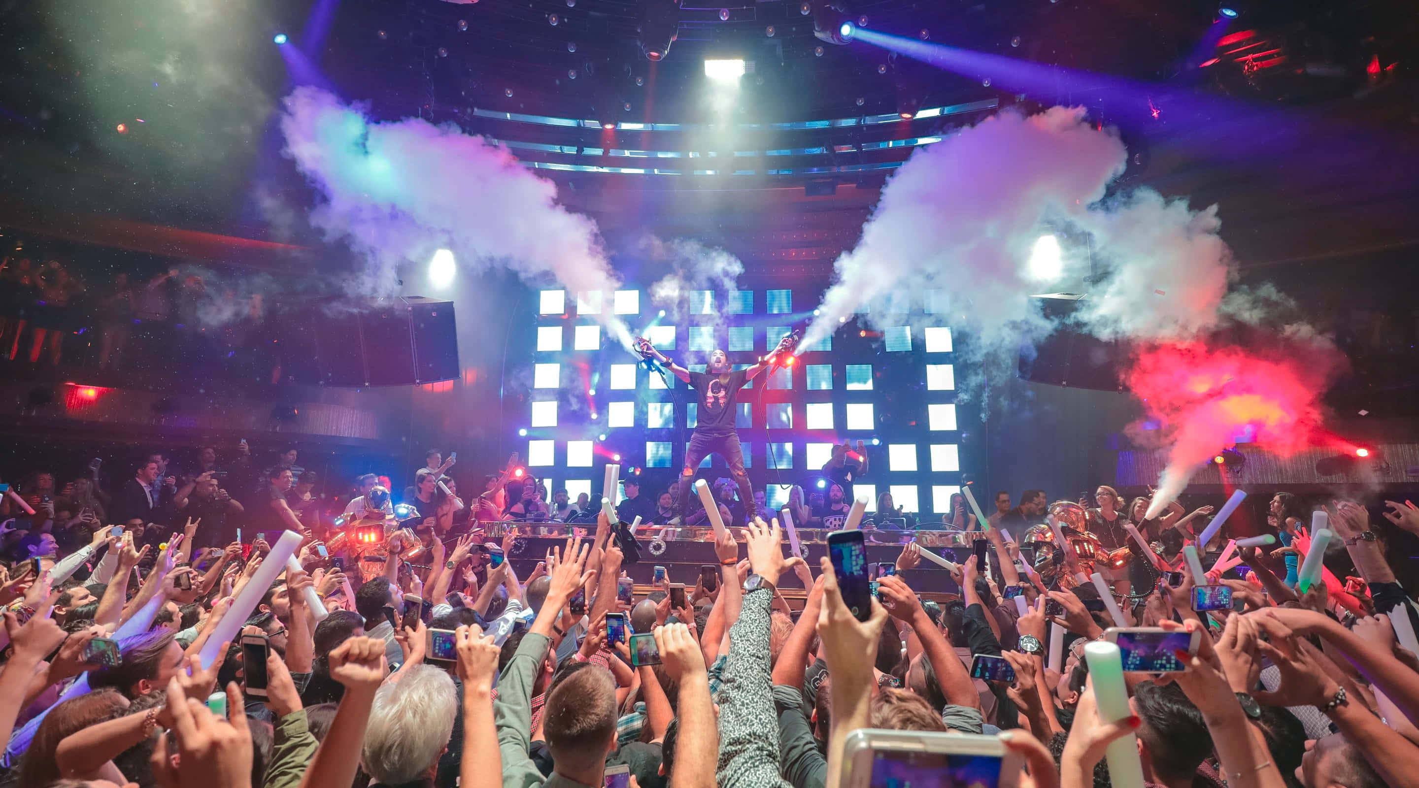 Nightclub 桌布 壁紙 — 表演者在舞台上跳躍，周圍煙霧繚繞、燈光璀璨，狂歡的人群高舉手機和螢光棒。