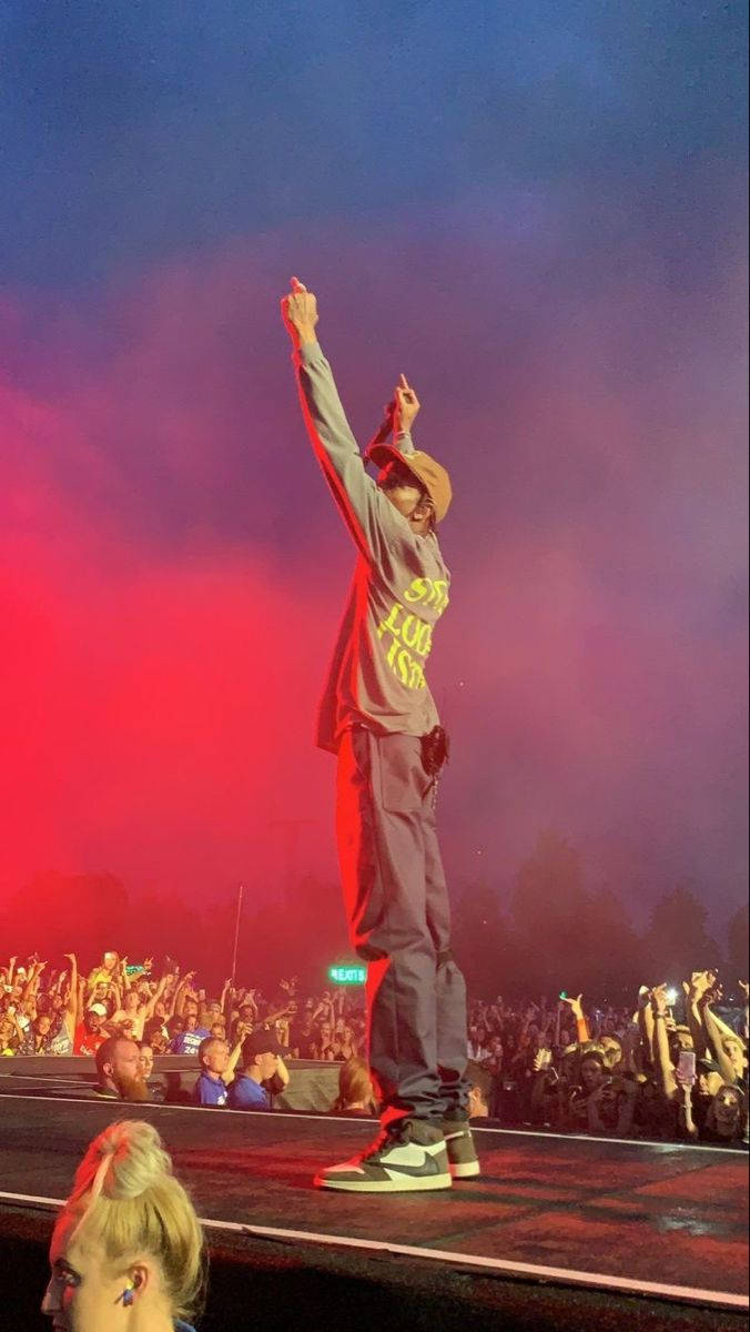Travis Scott Concert 桌布 壁紙 — 表演者在舞台上高舉雙臂，向在鮮豔紅色和藍色舞台燈光下歡呼的人群致意。