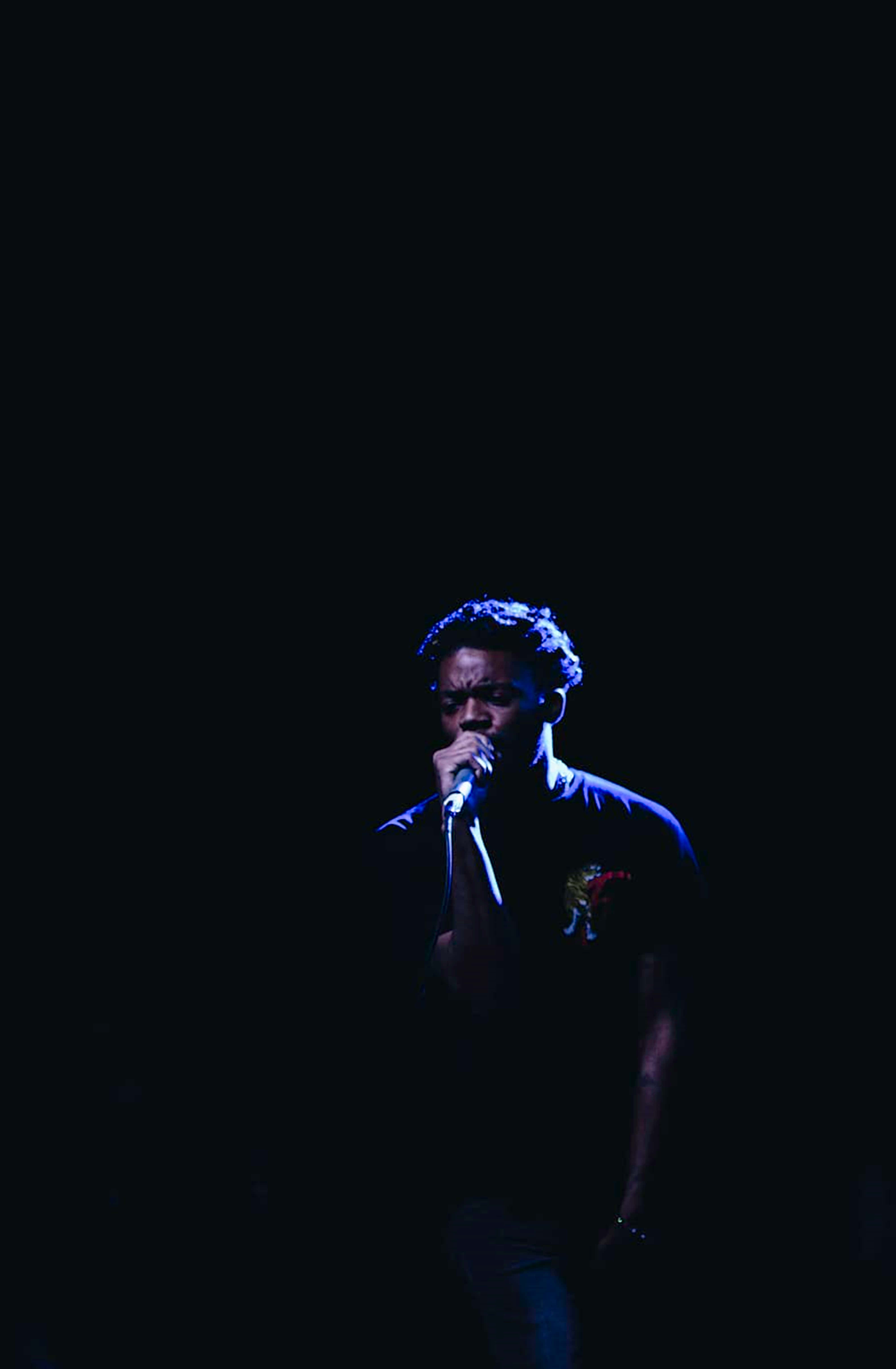 4K Rapper 桌布 壁紙 — 表演者在藍色舞台燈光照射下，對著漆黑背景向麥克風唱歌。
