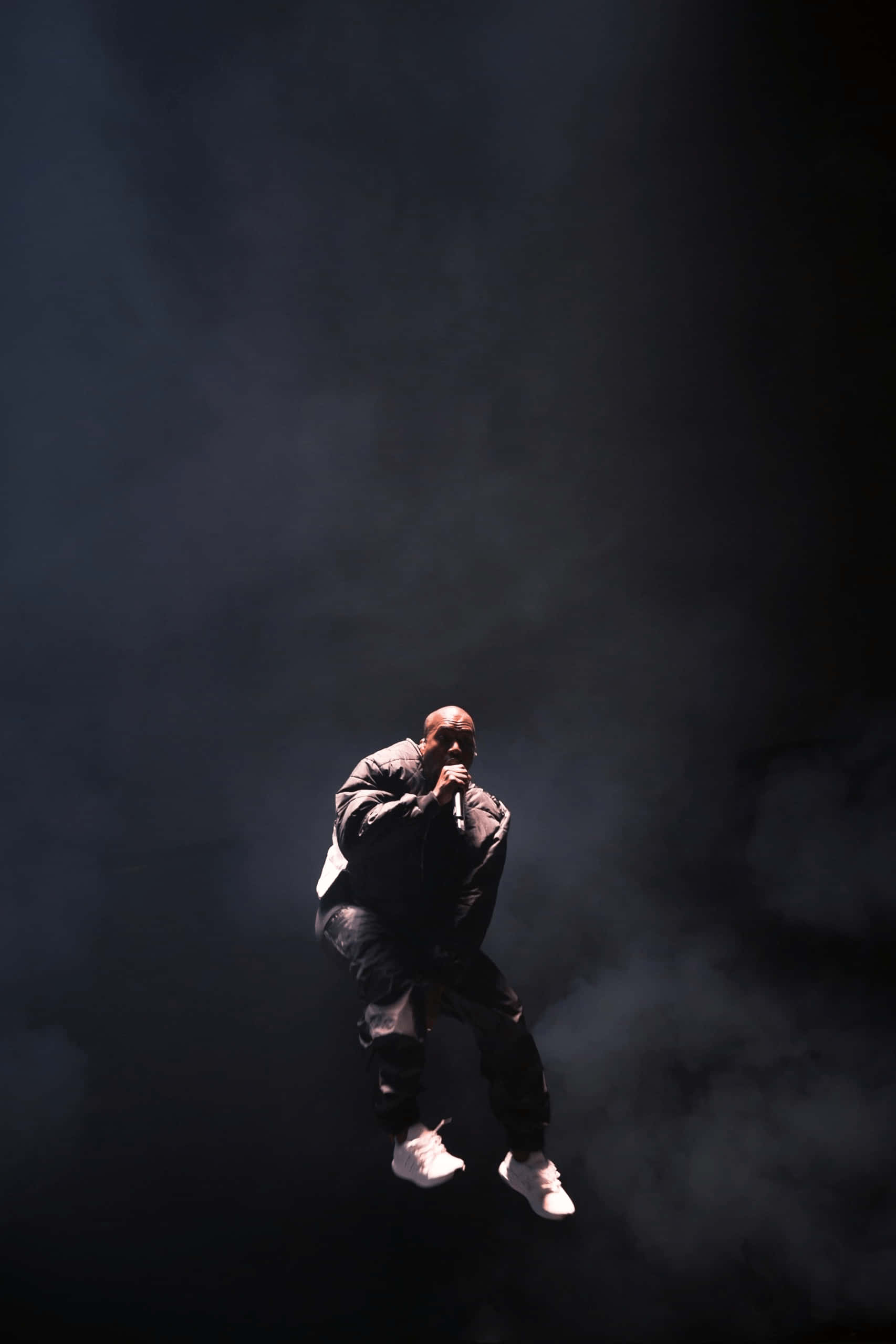 kanye iPhone 桌布 壁紙 — 表演者在黑暗煙霧中懸浮，在戲劇性舞台燈光下手持麥克風。