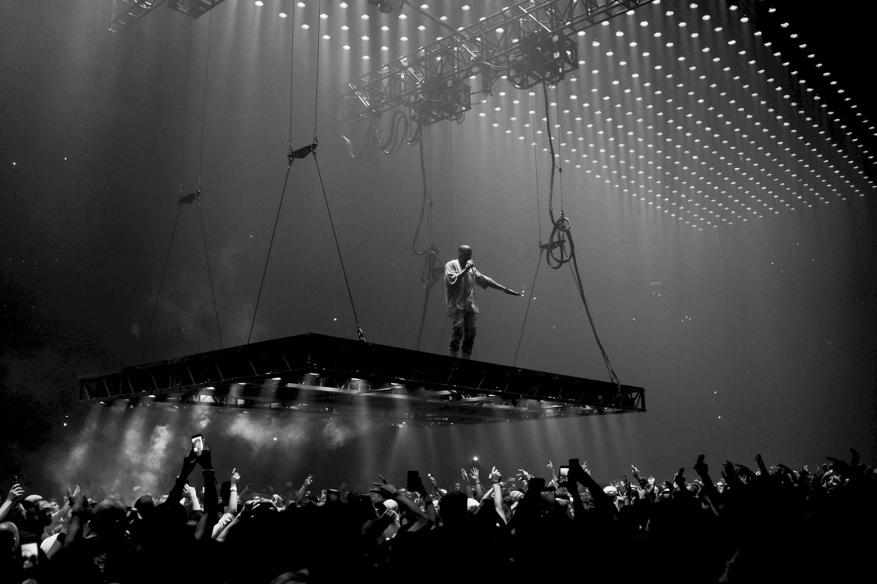Kanye 桌布 壁紙 — 表演者獨自站在懸掛的舞台上，下方是熱情歡呼的人群，在戲劇性的舞台燈光下。
