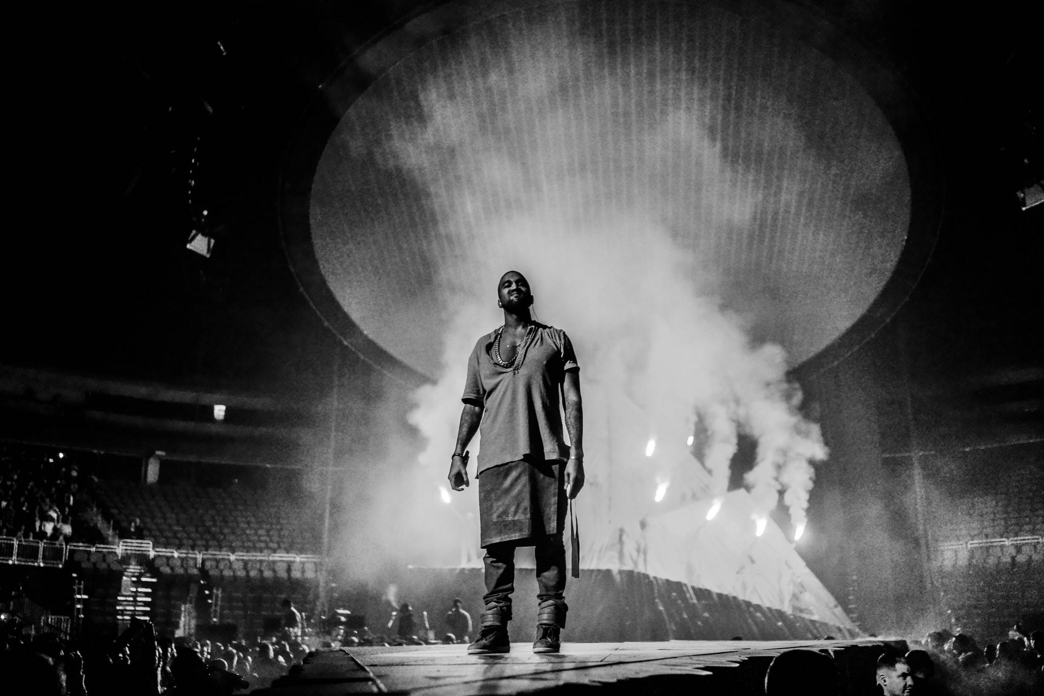 Kanye West Donda 桌布 壁紙 — 表演者獨自站在煙霧瀰漫的音樂會舞台上，在戲劇性的燈光下，觀眾在昏暗的場館中。