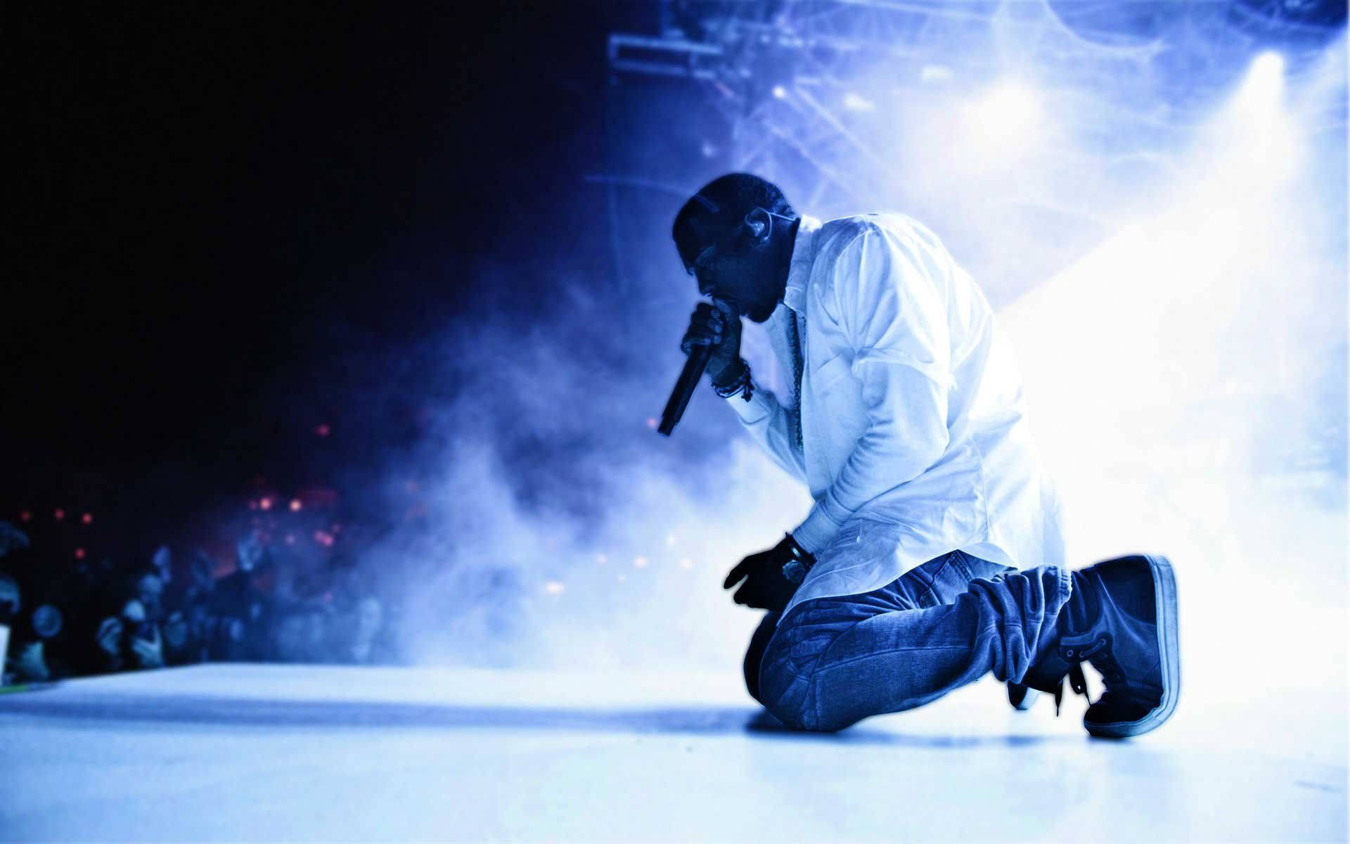 Kanye 桌布 壁紙 — 表演者跪在舞台上，沐浴在藍光中，手持麥克風，煙霧在他周圍旋轉。