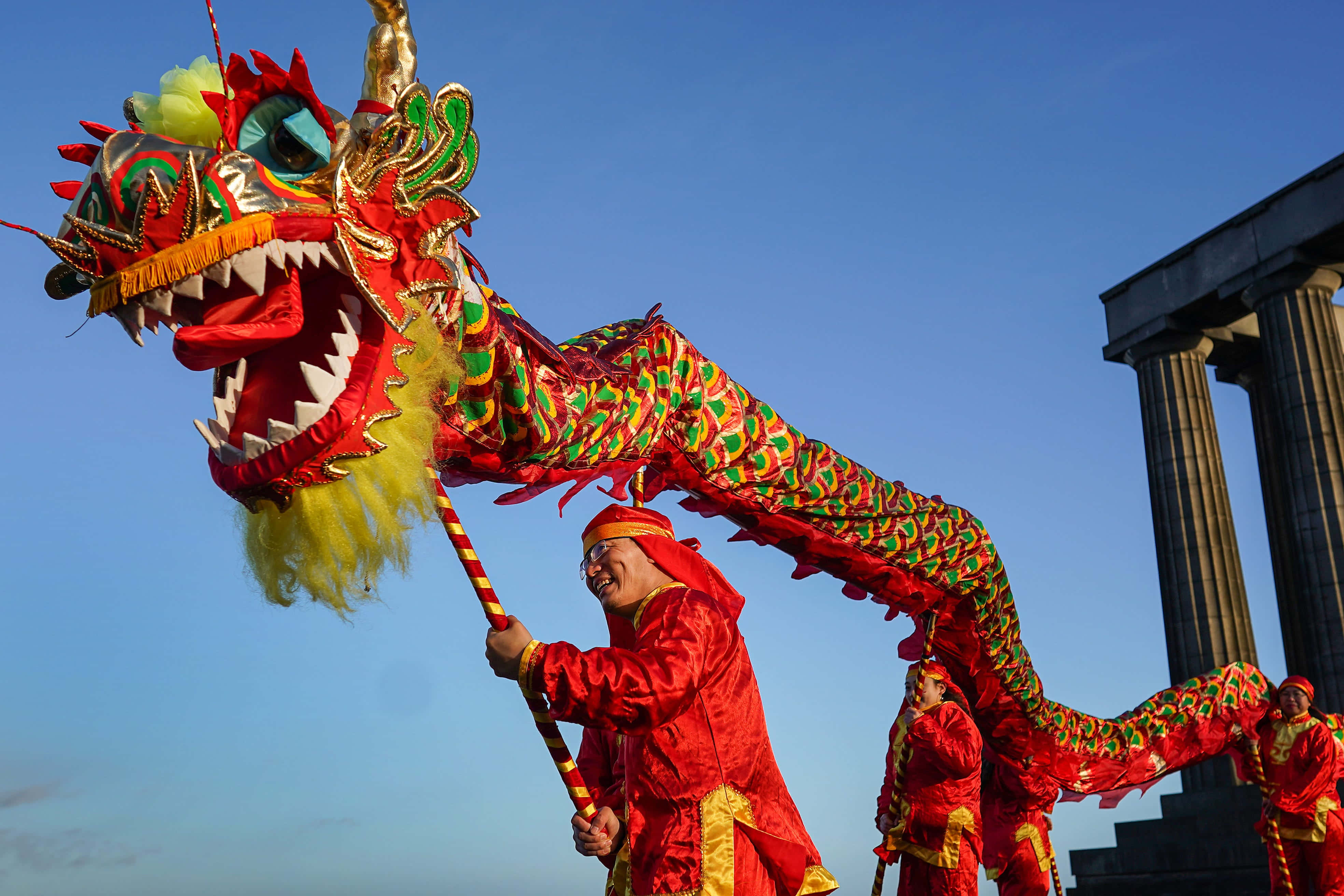 New Year 桌布 壁紙 — 表演者身穿紅色服裝，在古典柱子附近，藍天白雲下操控著鮮豔的紅綠龍偶。