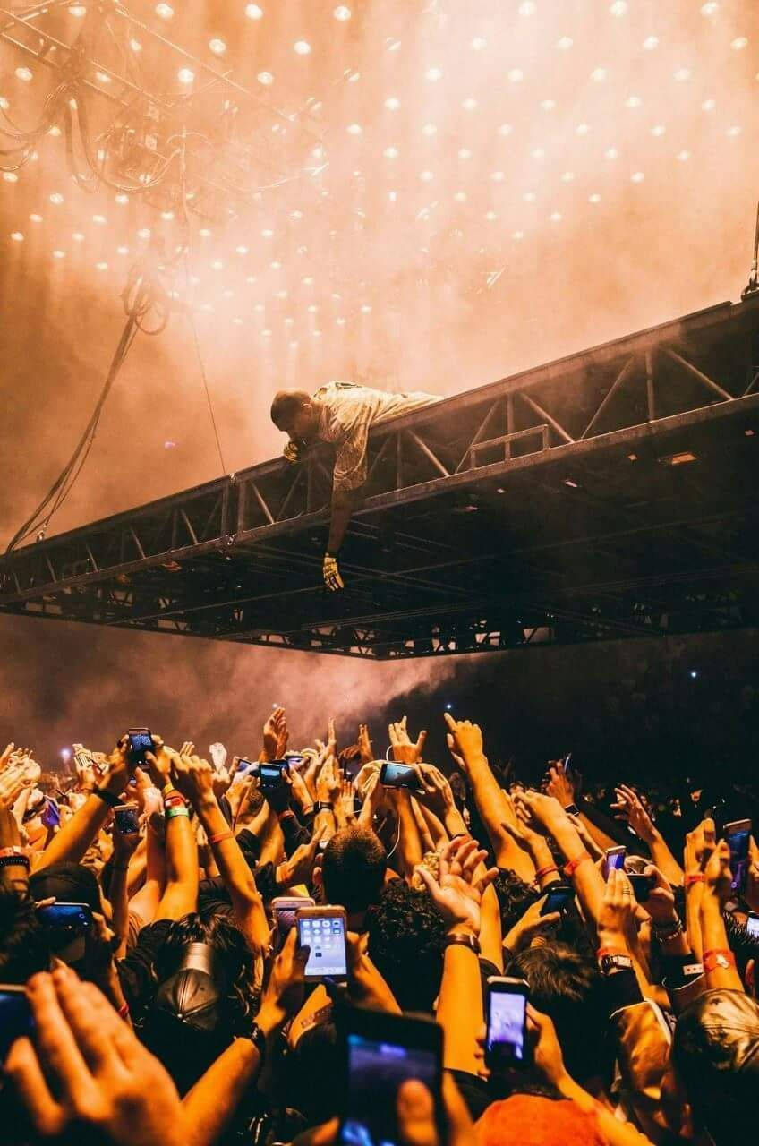 kanye west saint pablo 桌布 壁紙 — 表演者靠在舞台欄杆上，俯視著一片高舉的手和發光的手機螢幕，沐浴在溫暖的橙色舞台燈光中。