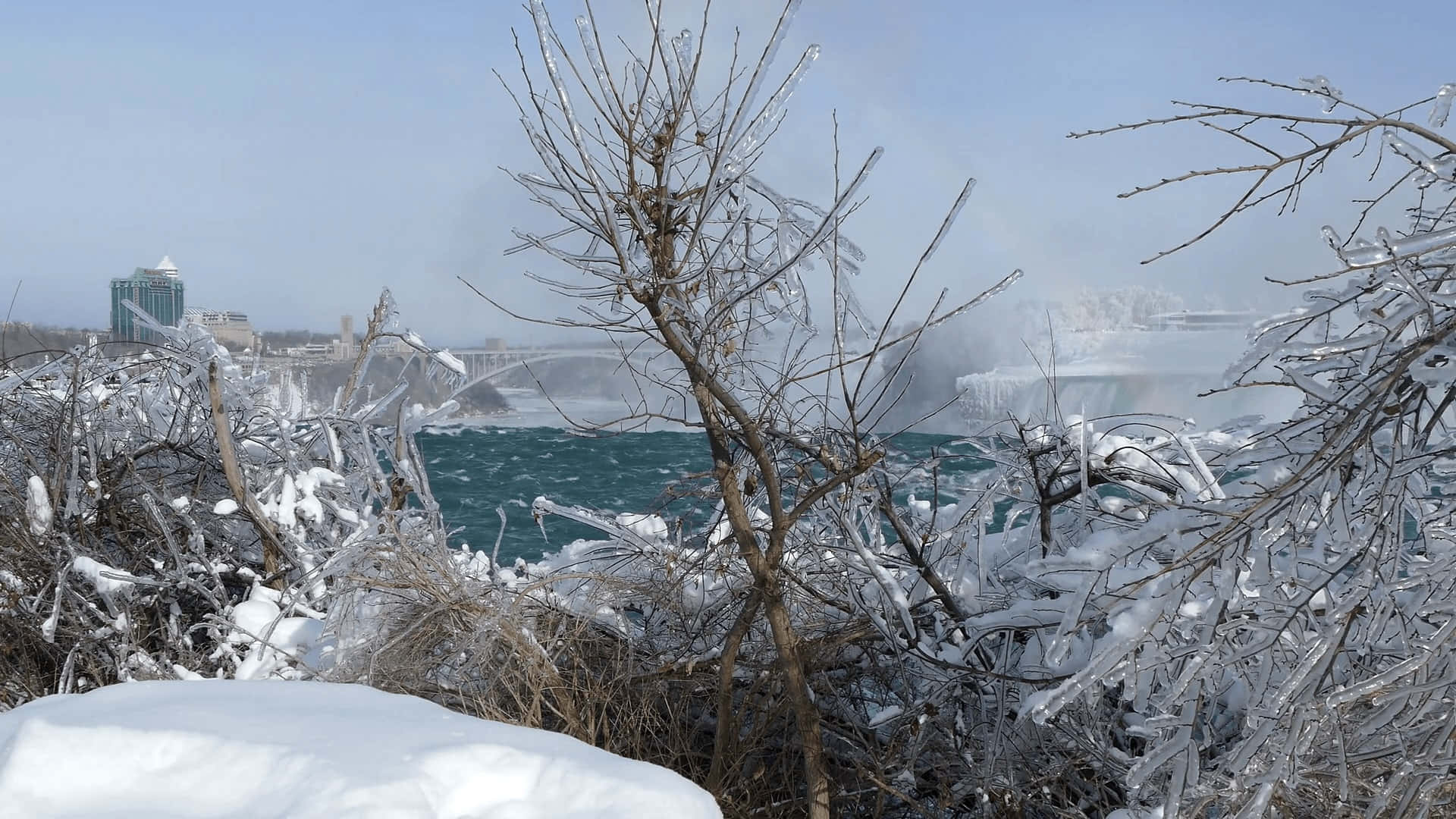 Polar Vortex 桌布 壁紙 — 被冰覆蓋的冰凍樹枝框住了尼亞加拉瀑布的景色，瀑布部分結冰，湍急的水面上升起薄霧。