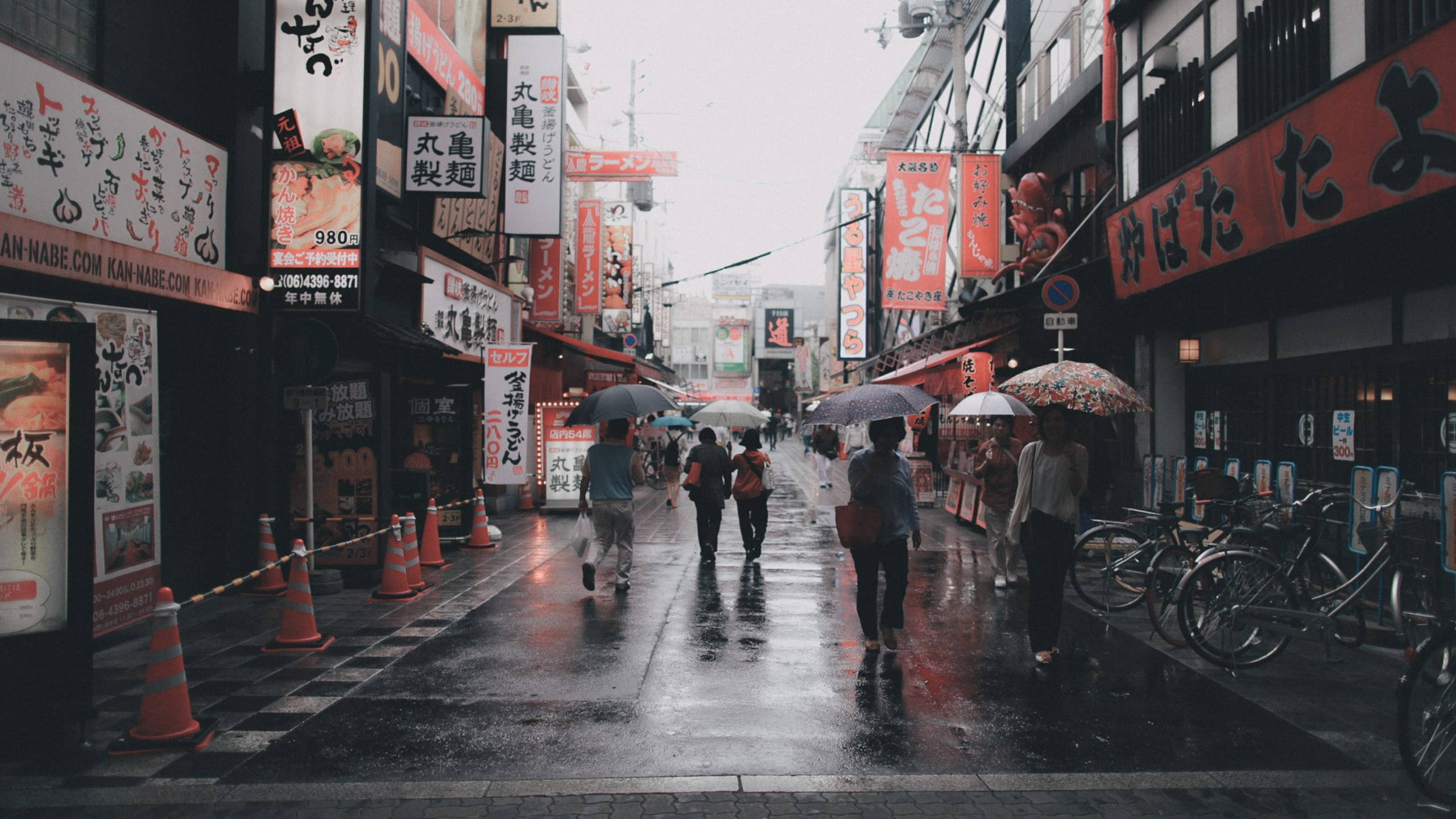 日本 桌布 壁紙 — 被雨水浸濕的日本小巷，行人撐著傘，在發光的霓虹招牌和掛燈下行走。