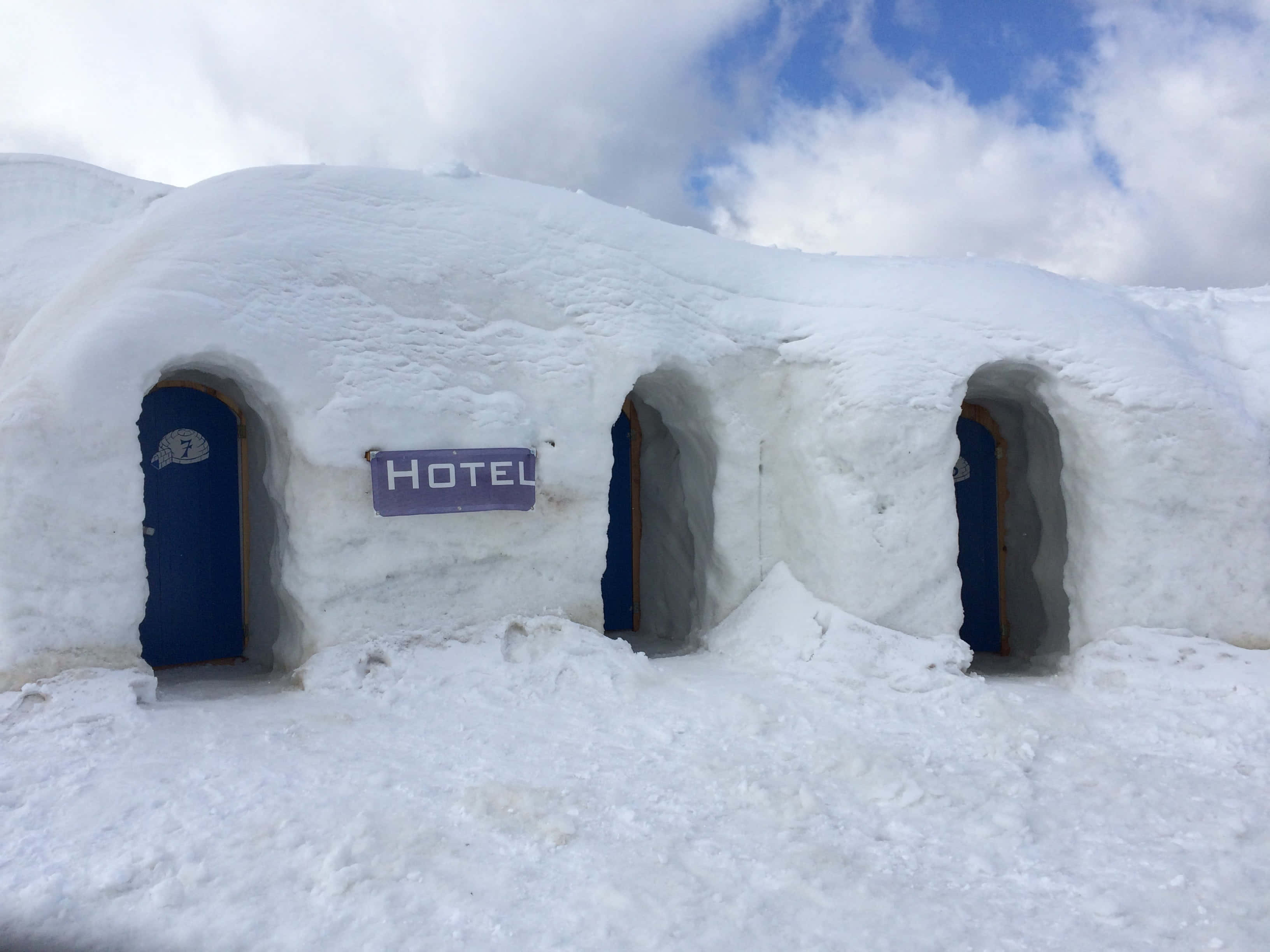 Ice Hotel 桌布 壁紙 — 被雪覆蓋的冰屋酒店，有藍色門和招牌，在陰天天空下。