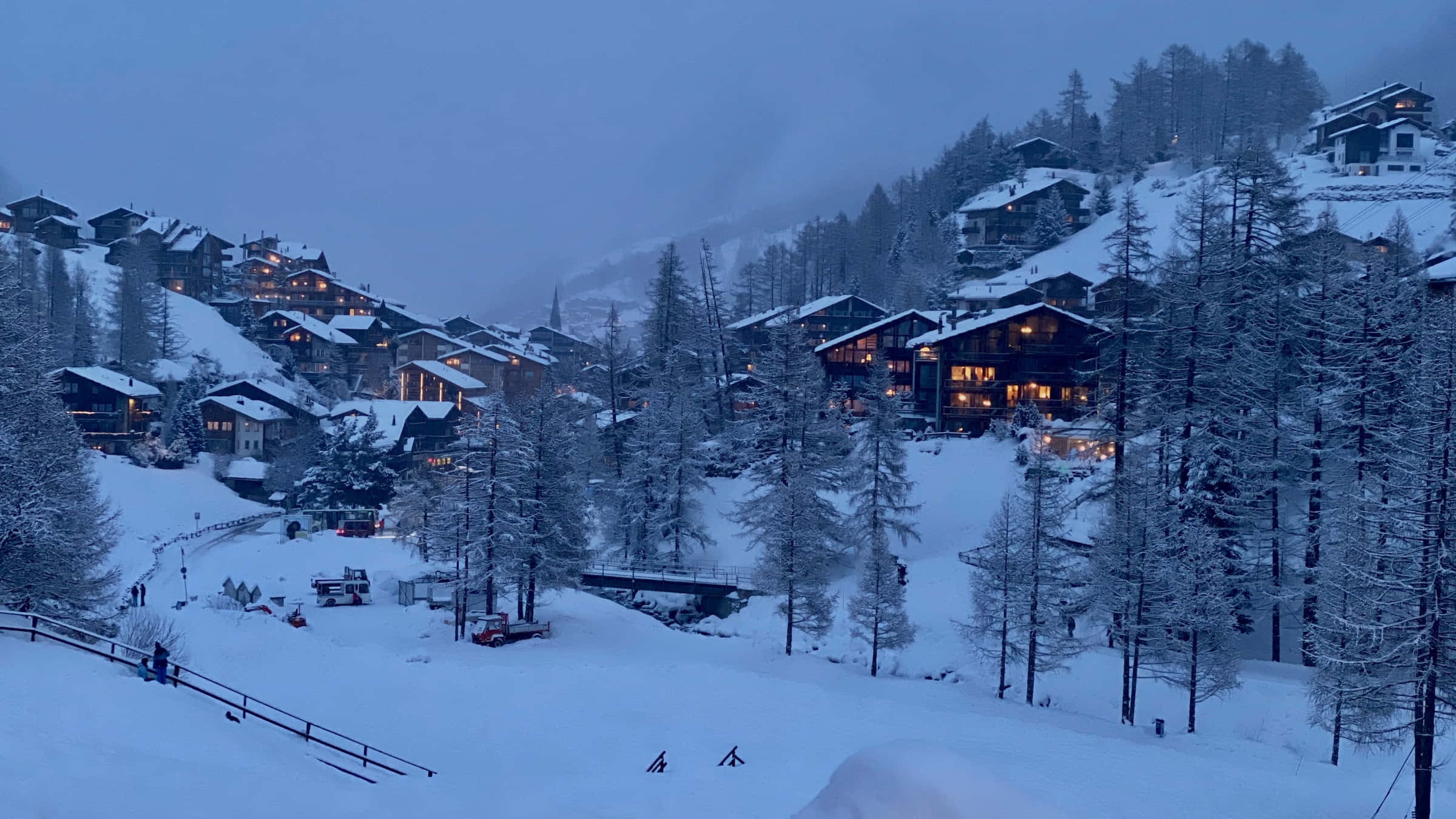 Ski Resort 桌布 壁紙 — 被雪覆蓋的小木屋在霧氣瀰漫的黃昏天空中溫暖地發光，坐落在松樹林中的山村裡。