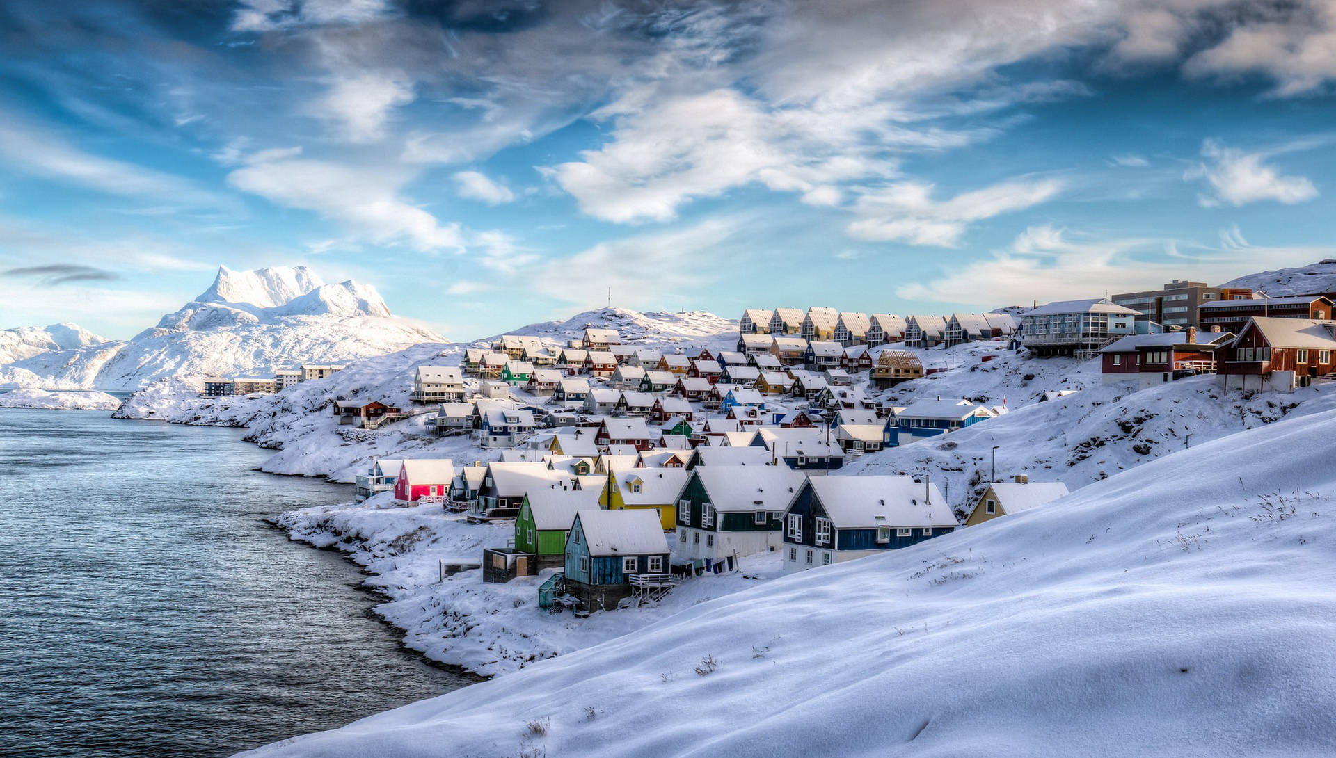 Greenland 桌布 壁紙 — 被雪覆蓋的彩色房屋依附在積雪的山坡上，旁邊是平靜的峽灣，上方是帶有縷縷白雲的明亮藍天。