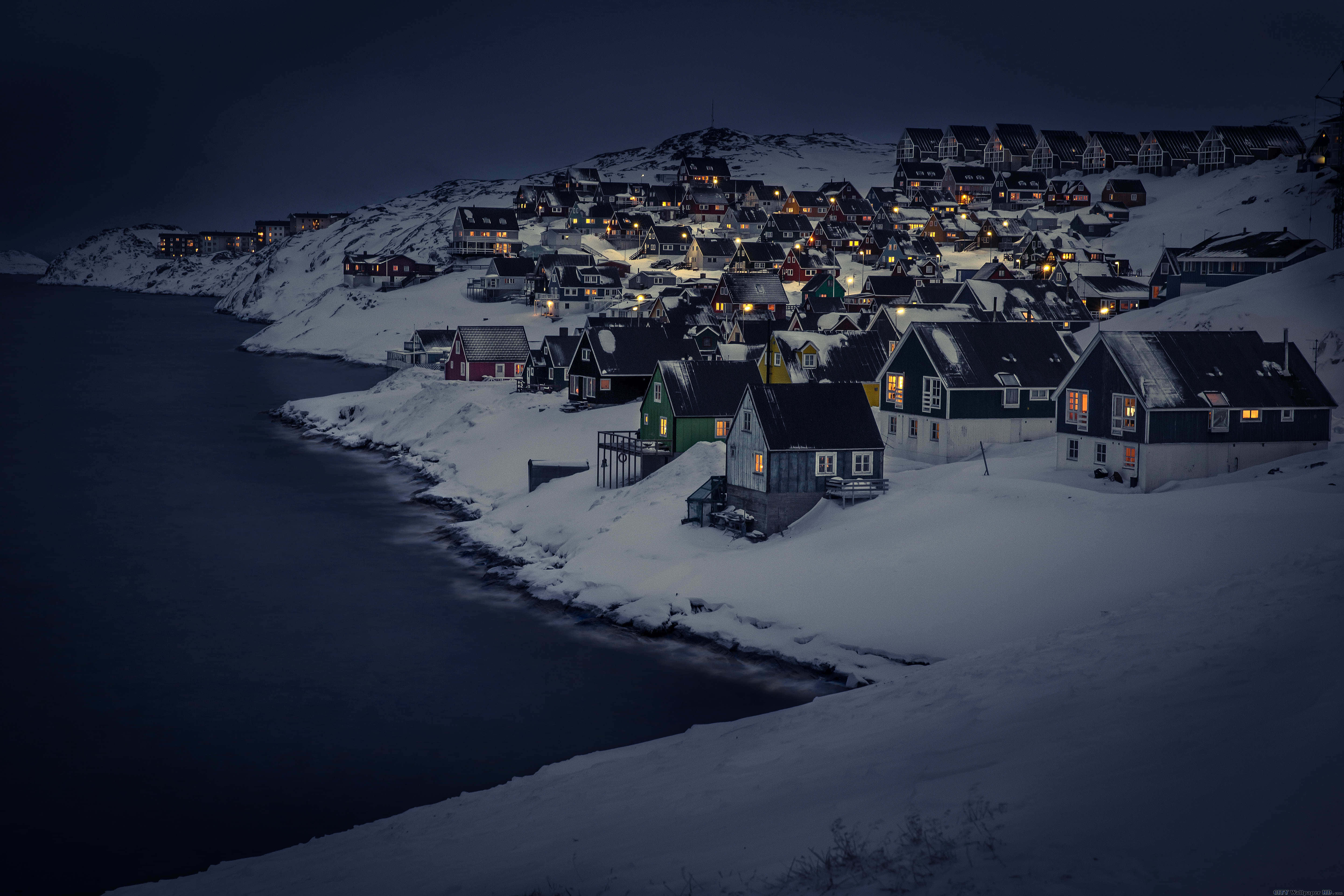 Greenland 桌布 壁紙 — 被雪覆蓋的房屋，窗戶閃爍著光芒，依附在黑暗的山坡上，旁邊是夜晚平靜的冰封峽灣。