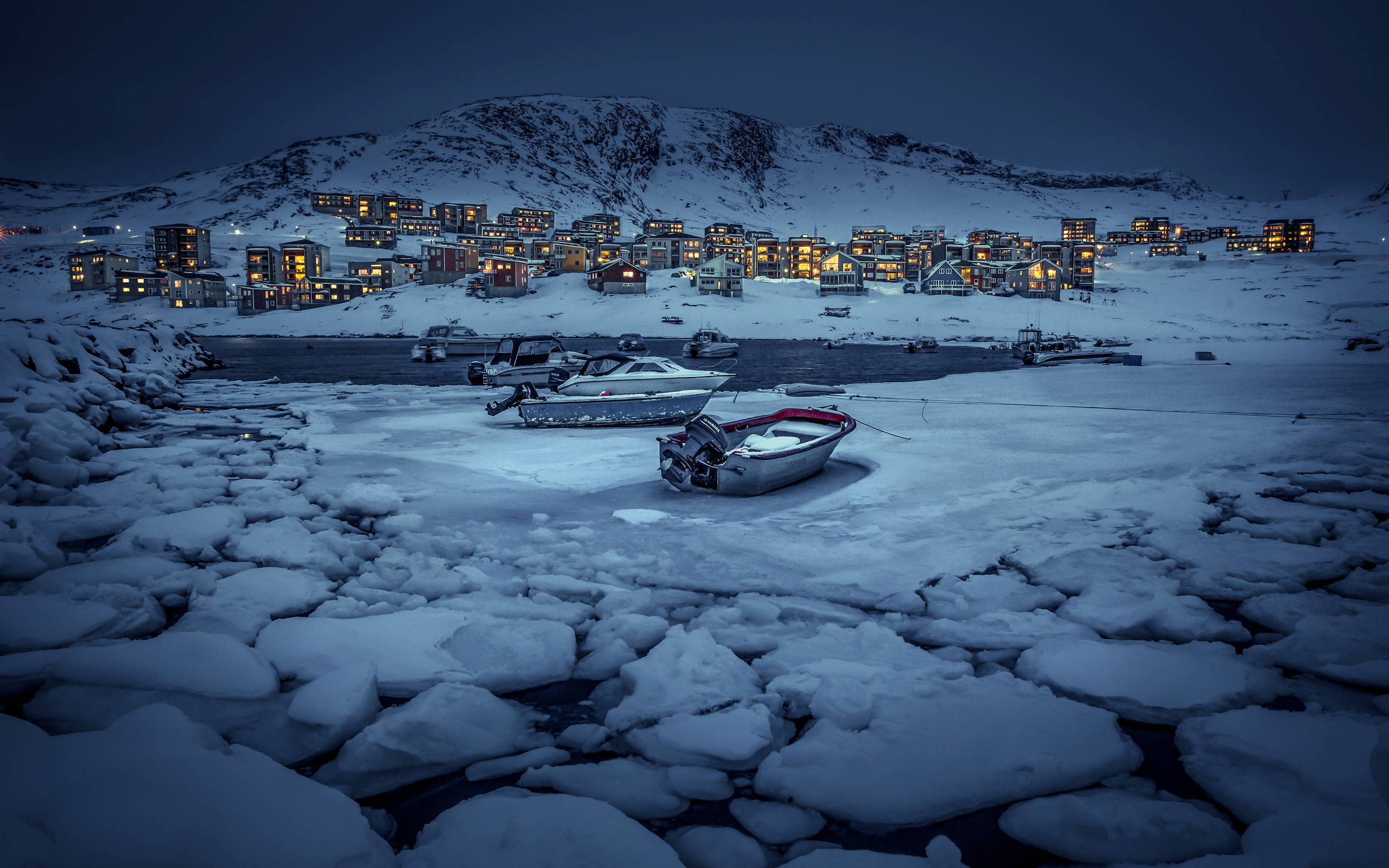 Greenland 桌布 壁紙 — 被雪覆蓋的船隻停泊在冰冷的水面上，溫暖的燈光從依附在雪坡上的房屋中在黃昏時分閃爍。