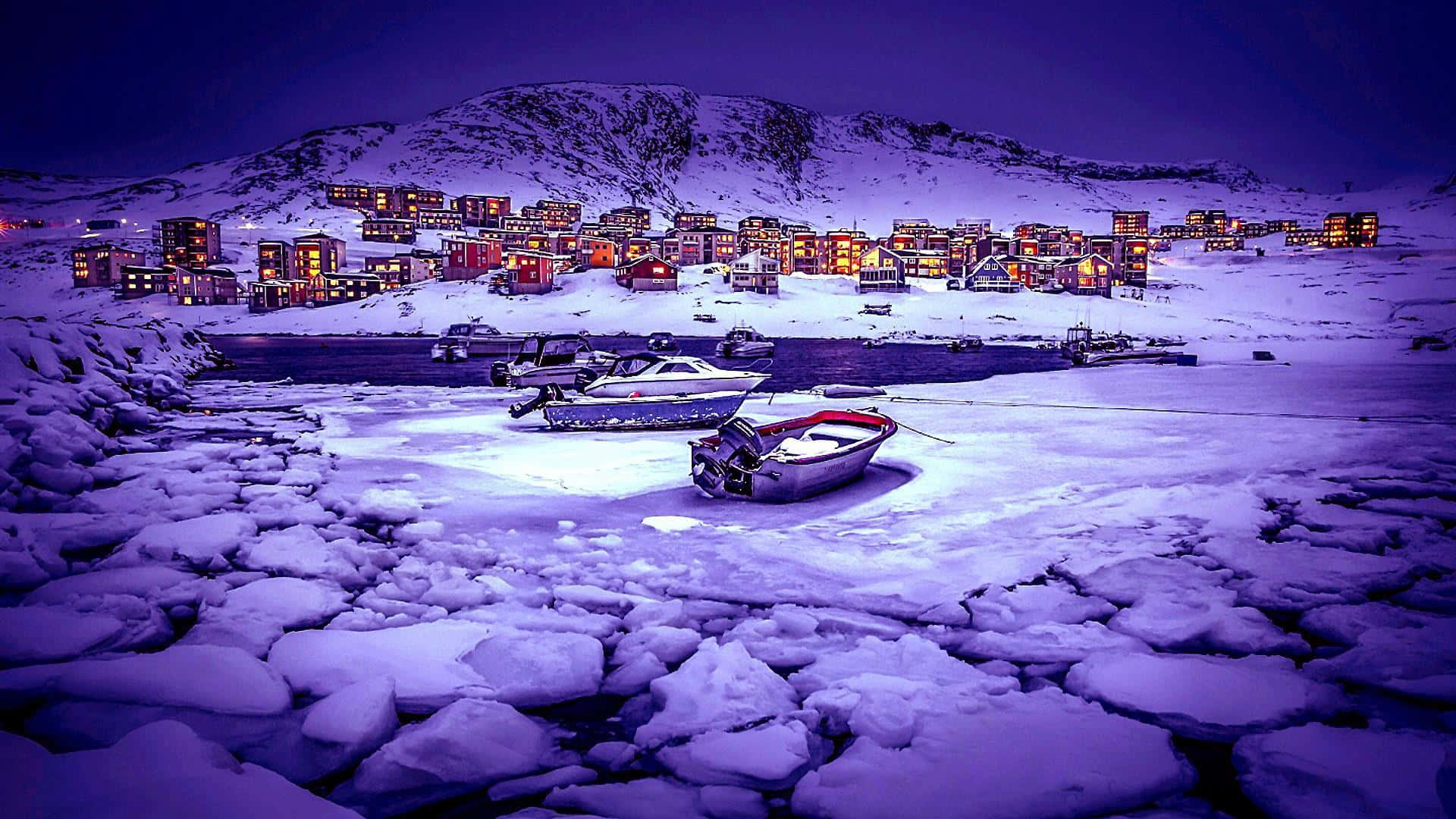 Greenland 桌布 壁紙 — 被雪覆蓋的船隻停泊在冰凍的水面上，前方是一個被雪覆蓋的山丘村莊，在深紫色的夜空下閃爍著溫暖的燈光。