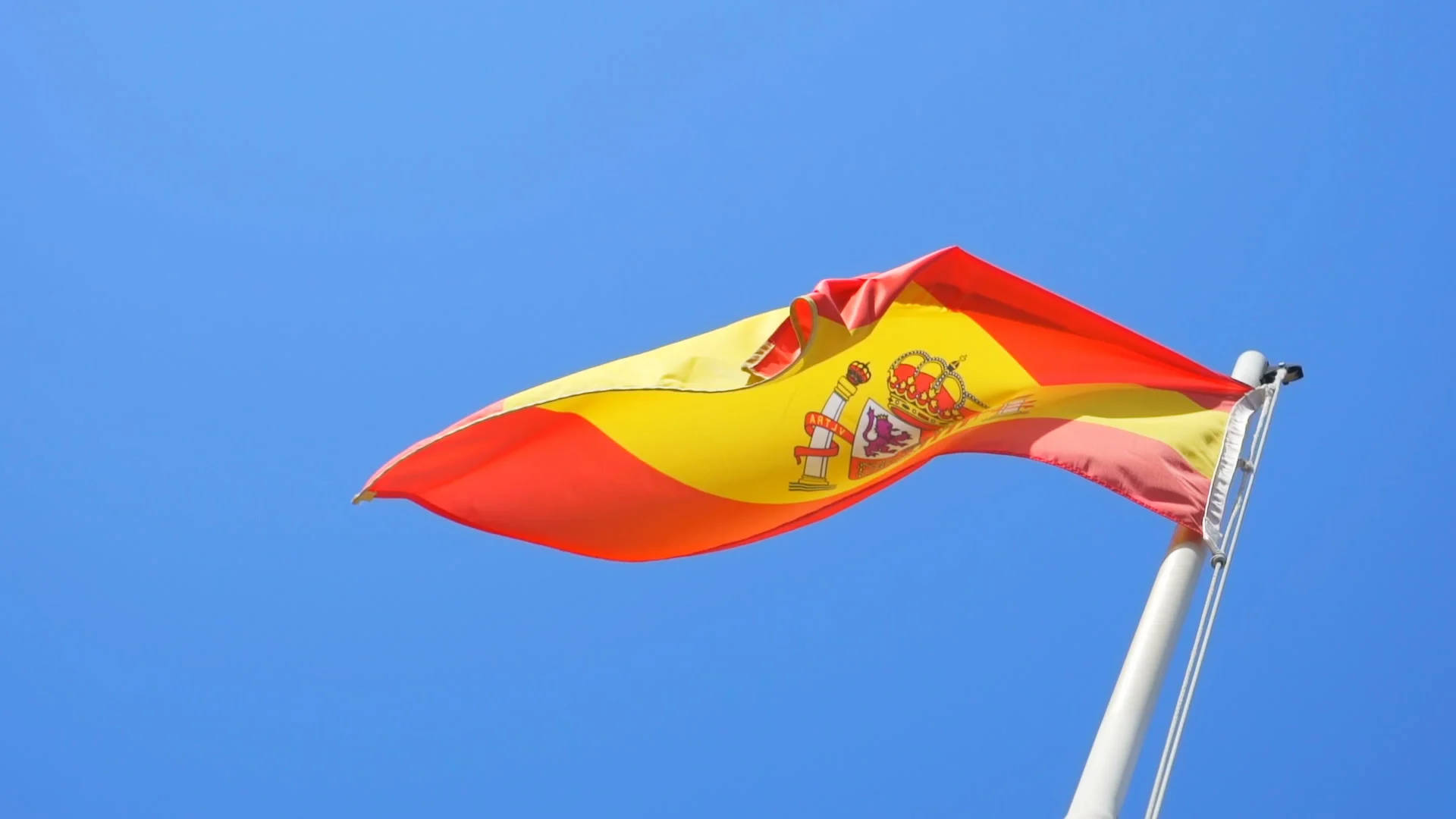 Spain Flag 桌布 壁紙 — 西班牙國旗，紅黃條紋和國徽，在清澈的藍天下大膽飄揚。
