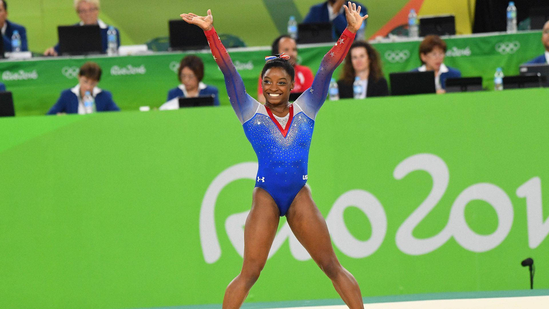 Simone Biles 桌布 壁紙 — 西蒙·拜爾斯在里約2016奧運會體操比賽後，身穿藍色閃亮緊身衣，高舉雙臂，勝利光芒四射。