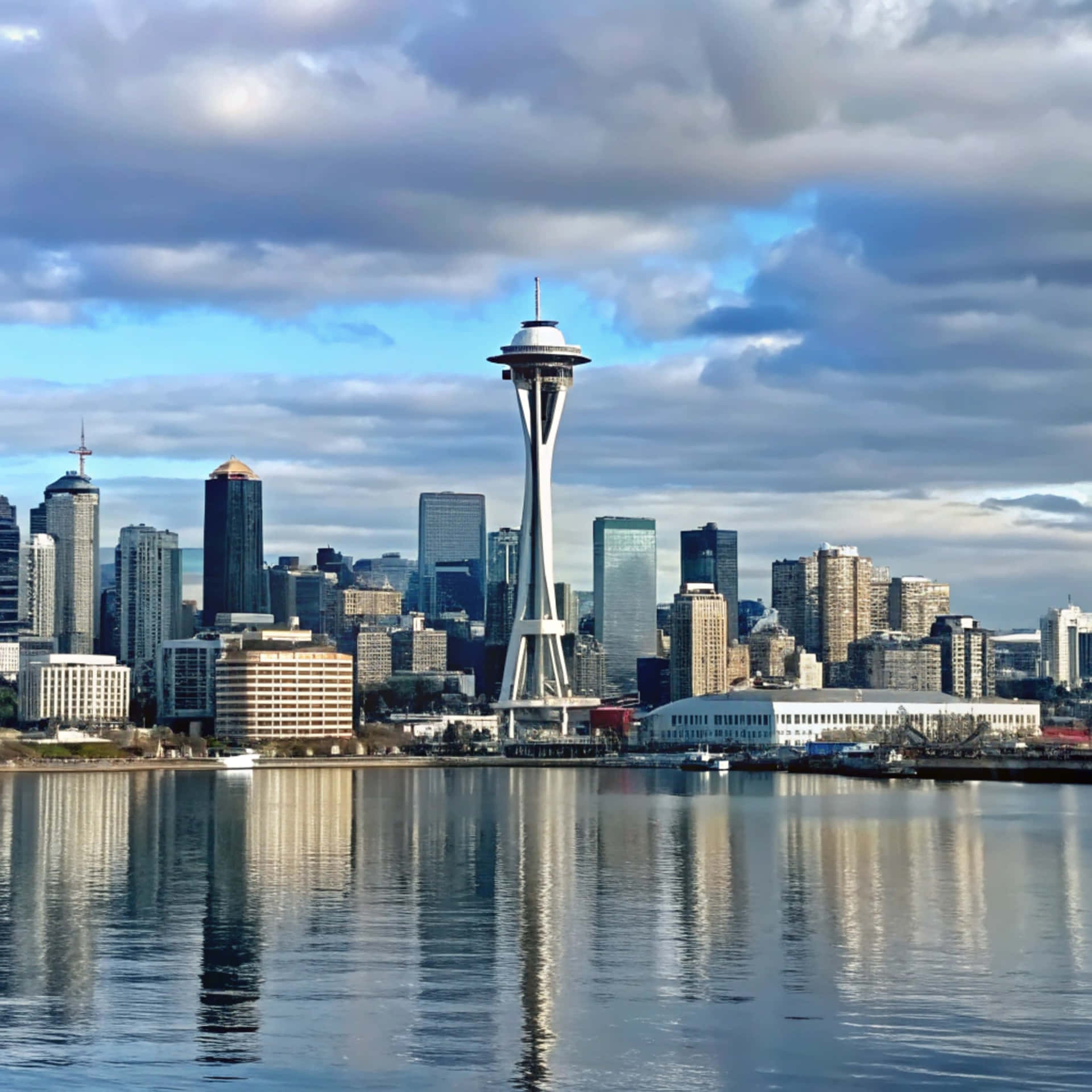 Seattle Skyline 壁紙 — 西雅圖天際線，太空針塔在戲劇性的多雲天空下聳立於海濱之上。