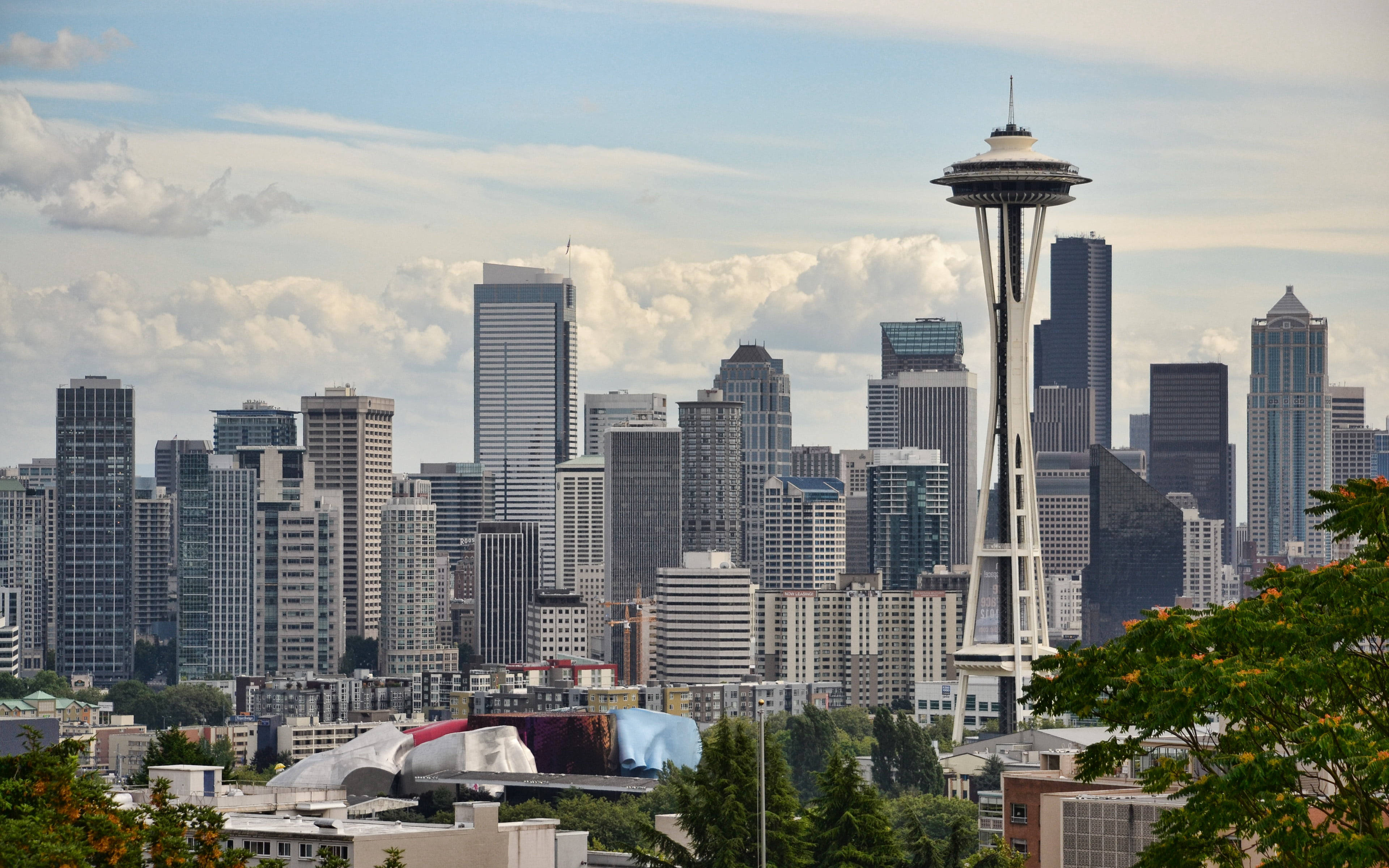 Space Needle 桌布 壁紙 — 西雅圖天際線，太空針塔聳立於玻璃摩天大樓之上，天空多雲，綠樹環繞。