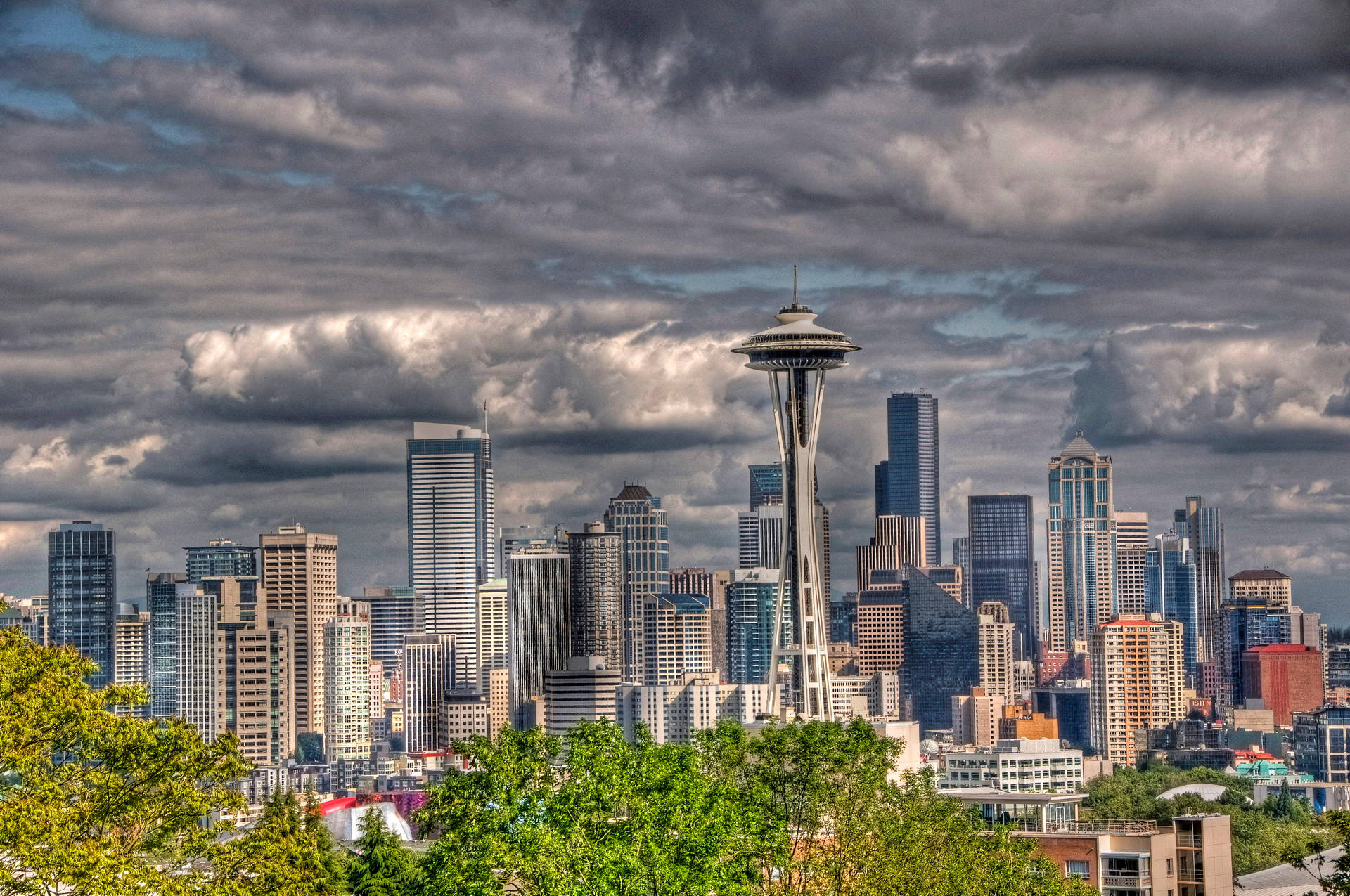 seattle 4K 桌布 壁紙 — 西雅圖天際線以太空針塔為主導，背景是戲劇性的暴風雲，前景是茂密的綠色樹木。