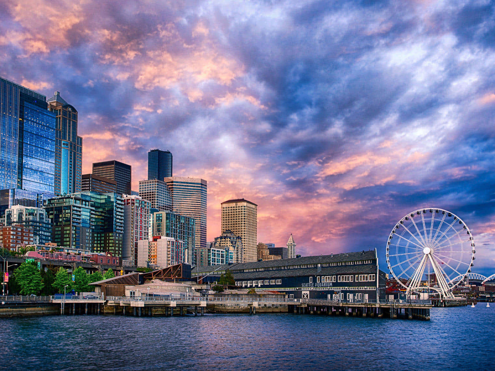 Best Seattle 桌布 壁紙 — 西雅圖天際線在日落時分，鮮豔的紫橙色雲彩倒映在西雅圖大摩天輪旁的水面上。