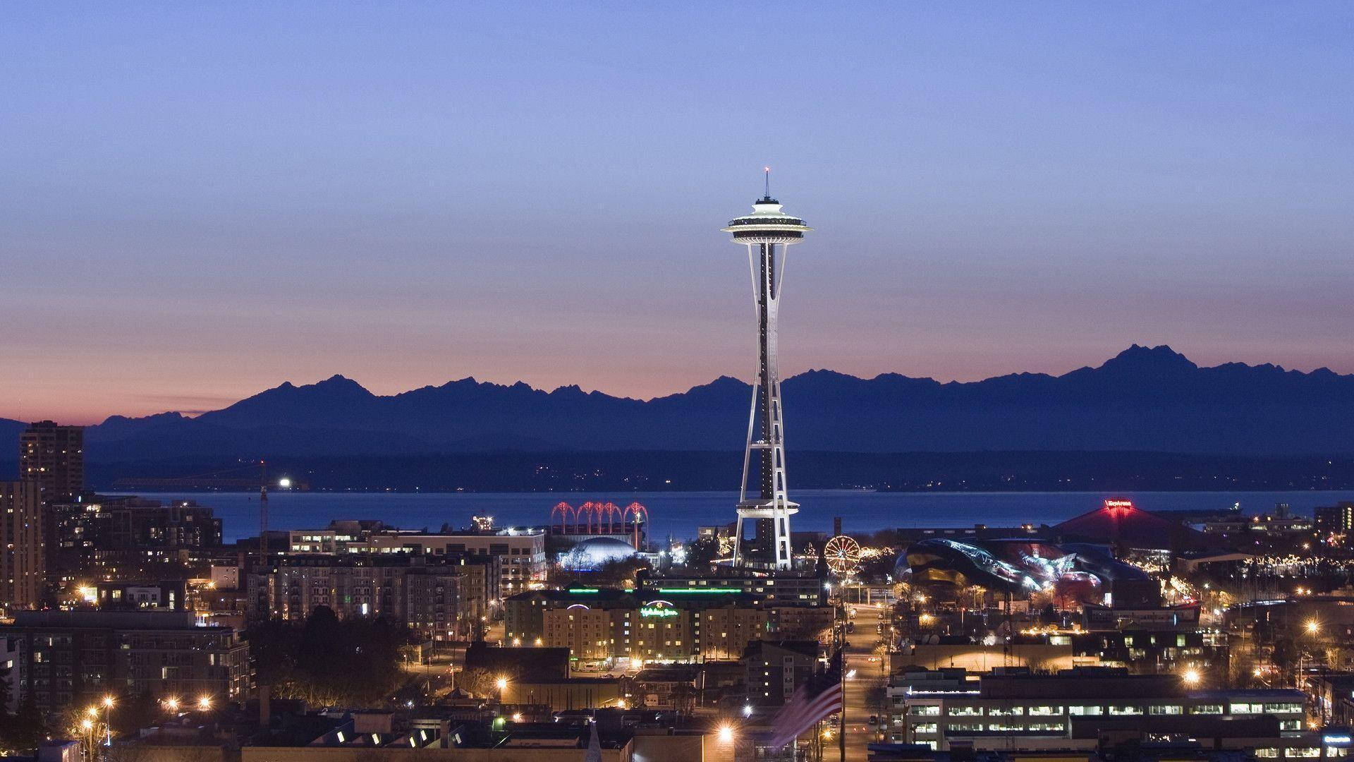 Space Needle 桌布 壁紙 — 西雅圖天際線在黃昏時分，太空針塔在紫色天空和遠山映襯下亮起燈光。