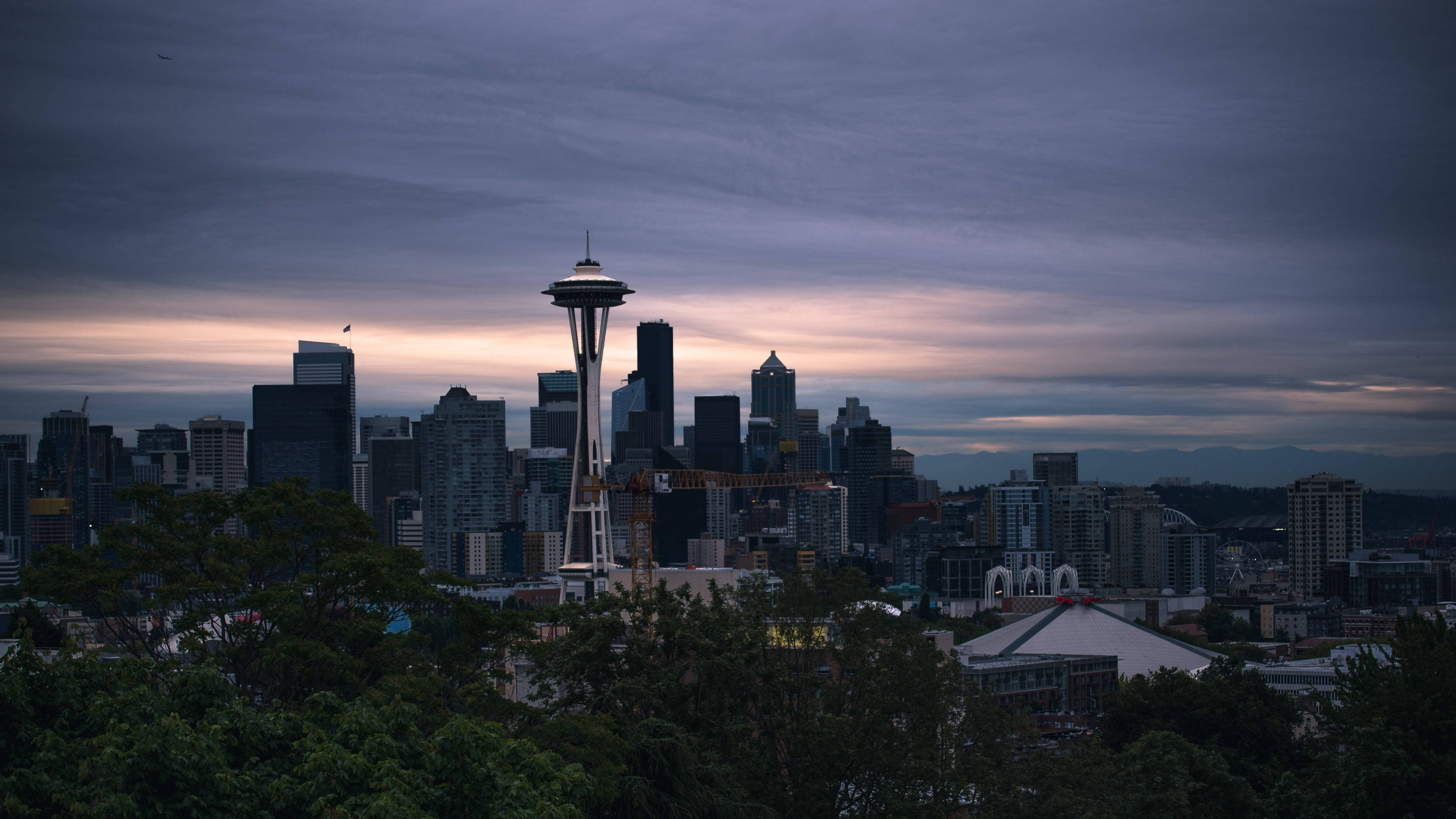Seattle Skyline 桌布 壁紙 — 西雅圖天際線在黃昏時分，太空針塔在陰鬱的紫色和橙色天空映襯下亮起，被黑暗的樹木框住。