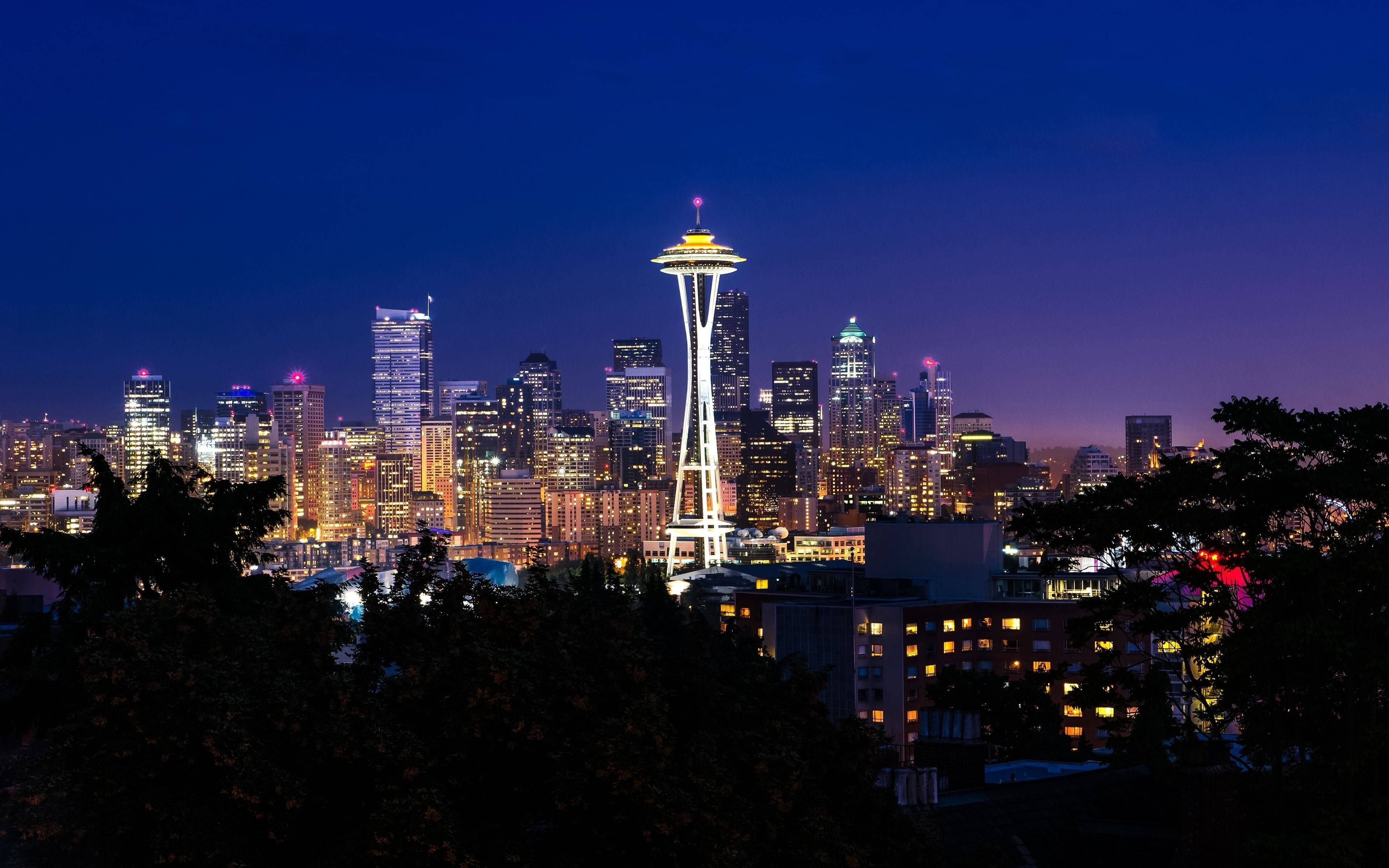 Seattle 桌布 壁紙 — 西雅圖天際線在黃昏時分，太空針塔在深藍色天空下閃耀，被黑暗的樹木剪影框住。