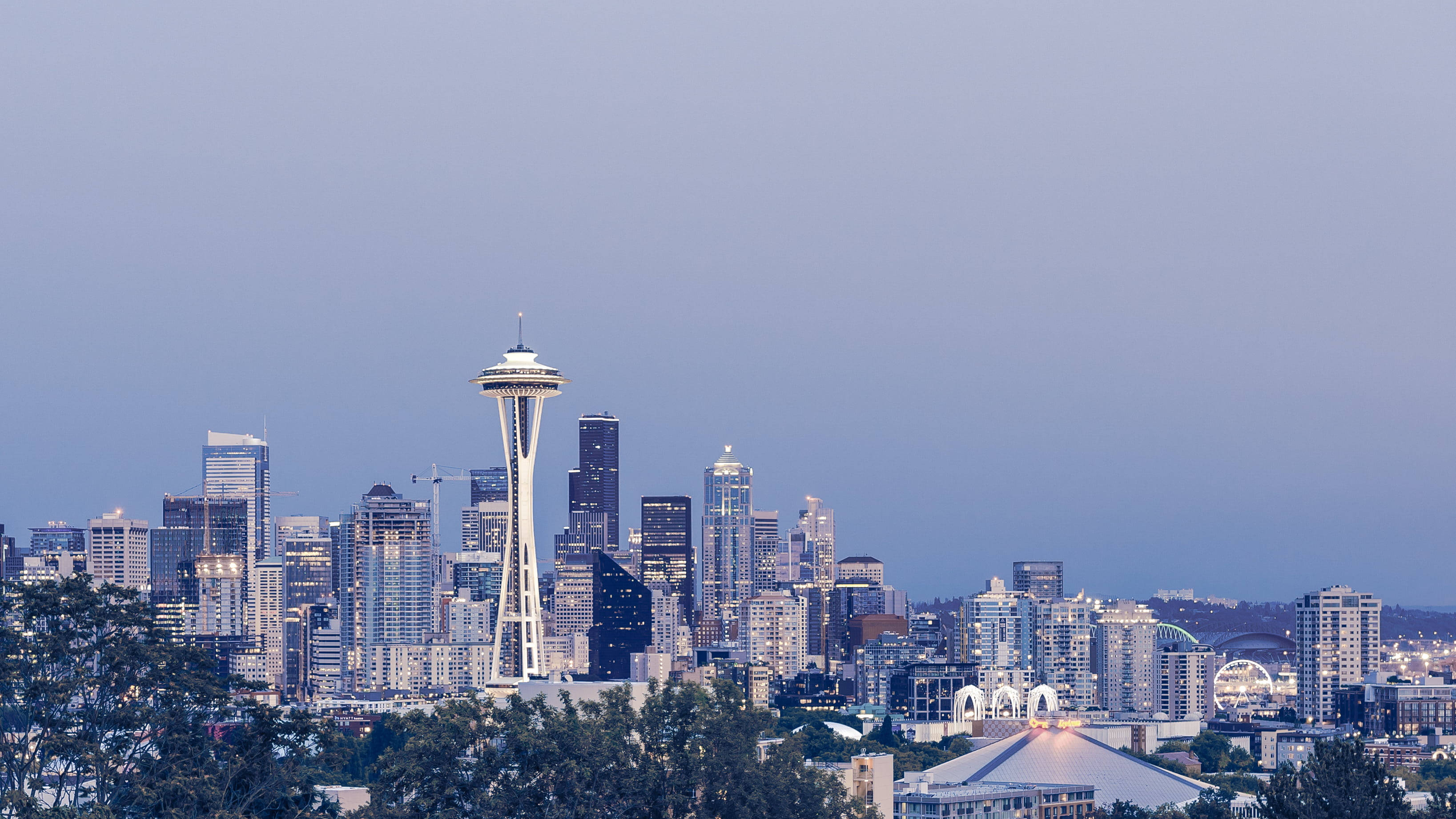 Space Needle 桌布 壁紙 — 西雅圖天際線在黃昏時分，太空針塔在柔和的紫色天空下亮起，前景有樹木剪影。