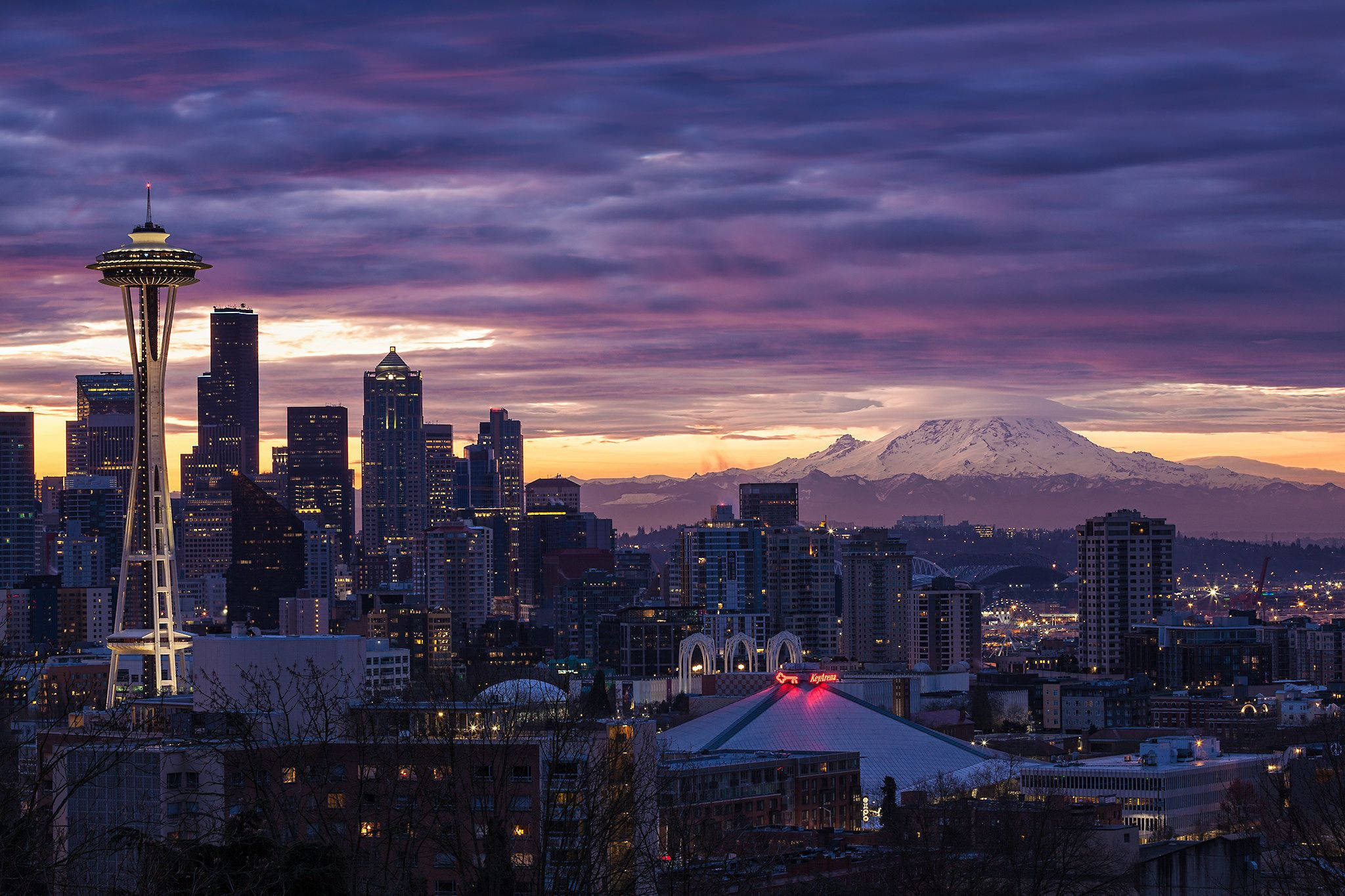 Seattle 桌布 壁紙 — 西雅圖天際線在黃昏時分，太空針塔在紫橙色天空下亮起，遠處是白雪覆蓋的雷尼爾山。