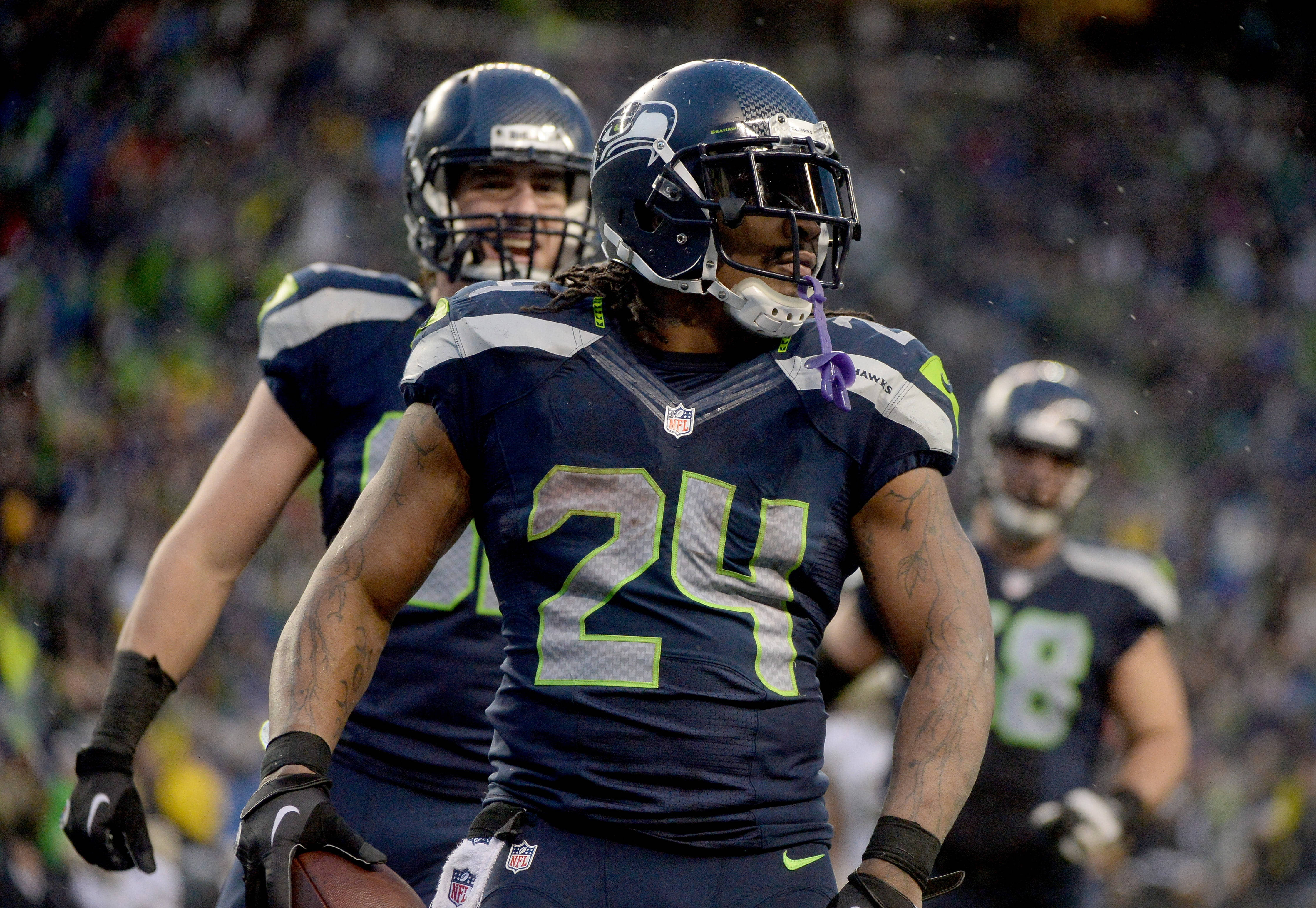 Seattle Seahawks 桌布 壁紙 — 西雅圖海鷹隊24號美式足球員在雨中比賽時嘴裡叼著紫色毛巾慶祝。