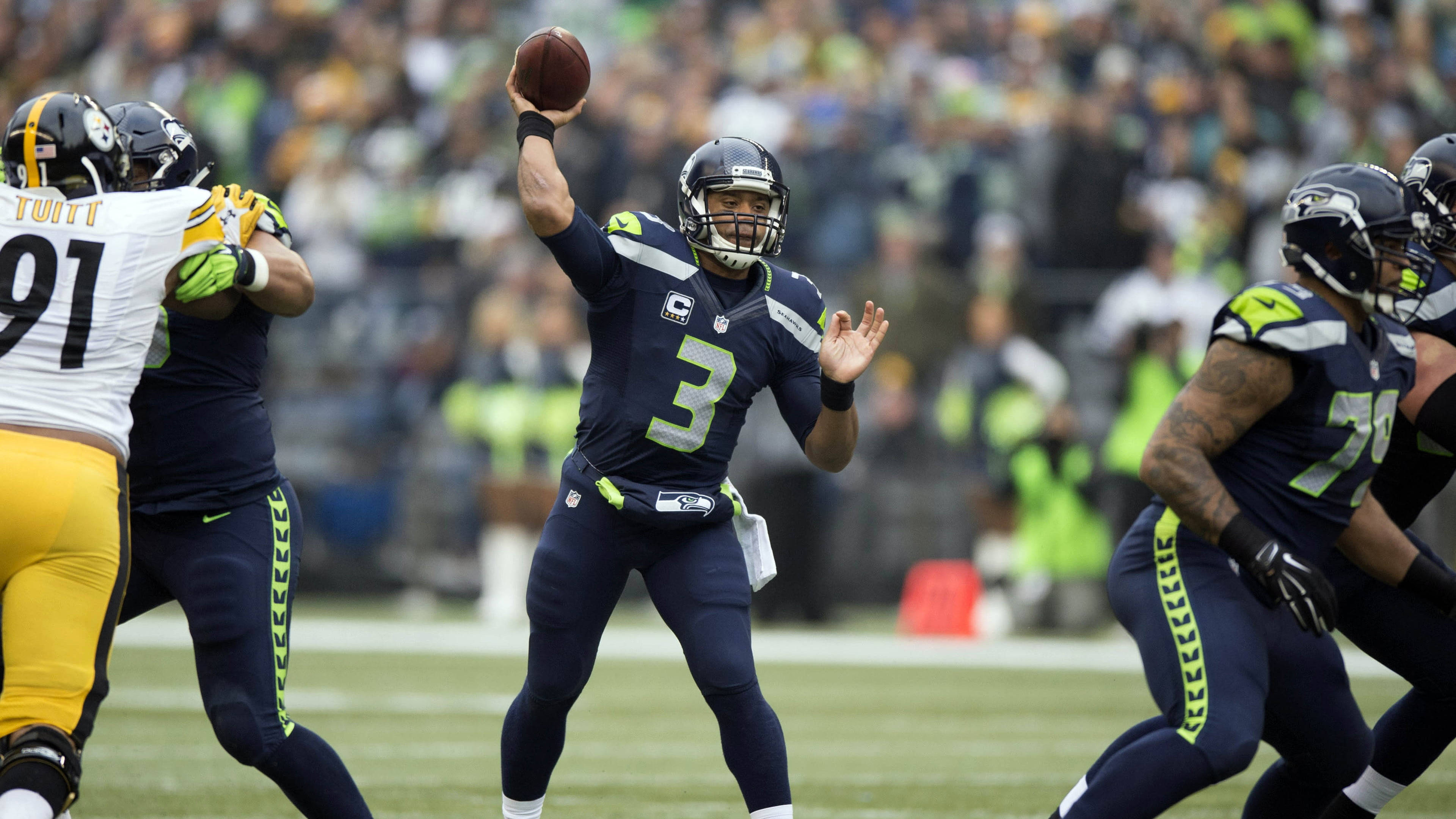 NFL Players 桌布 壁紙 — 西雅圖海鷹隊四分衛身穿海軍藍制服，在NFL日間比賽中擲出橄欖球，背景是模糊的人群。