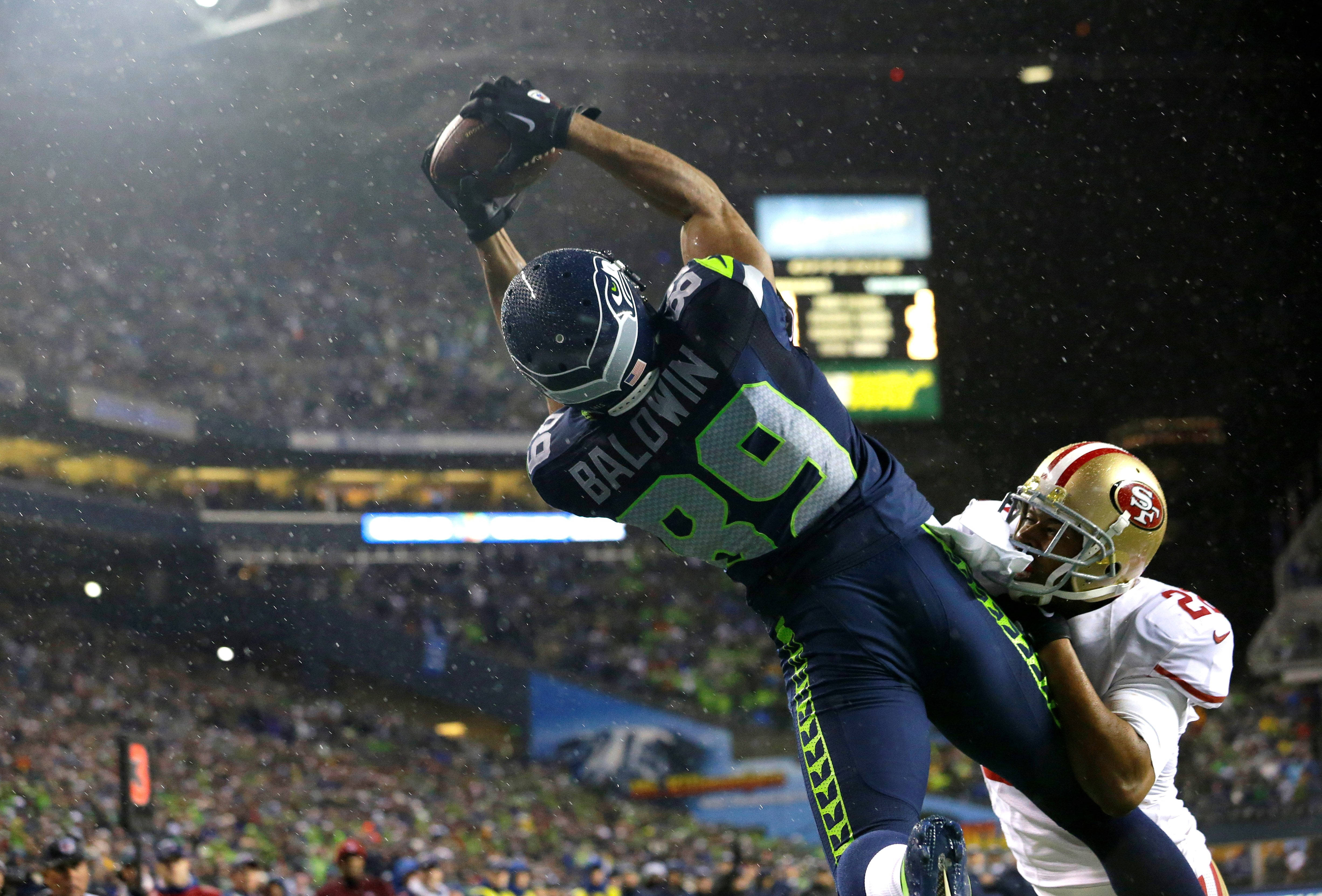 Seattle Seahawks 桌布 壁紙 — 西雅圖海鷹隊球員跳起接住美式足球，而舊金山49人隊防守球員在濕透的球場上擒抱他。