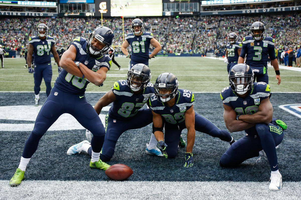 Seattle Seahawks 桌布 壁紙 — 西雅圖海鷹隊球員身穿海軍藍色制服，在比賽期間自信地在球場上擺姿勢，腳邊放著一個足球。