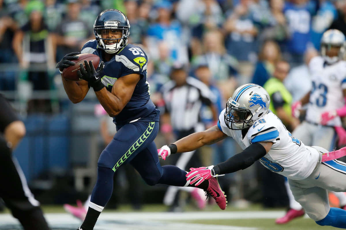 Fantasy Football Profile 桌布 壁紙 — 西雅圖海鷹隊的一名接球手身穿藍色球衣，帶著橄欖球奔跑，而底特律雄獅隊的一名白色球衣防守球員在陽光明媚的球場上撲向他進行擒抱。
