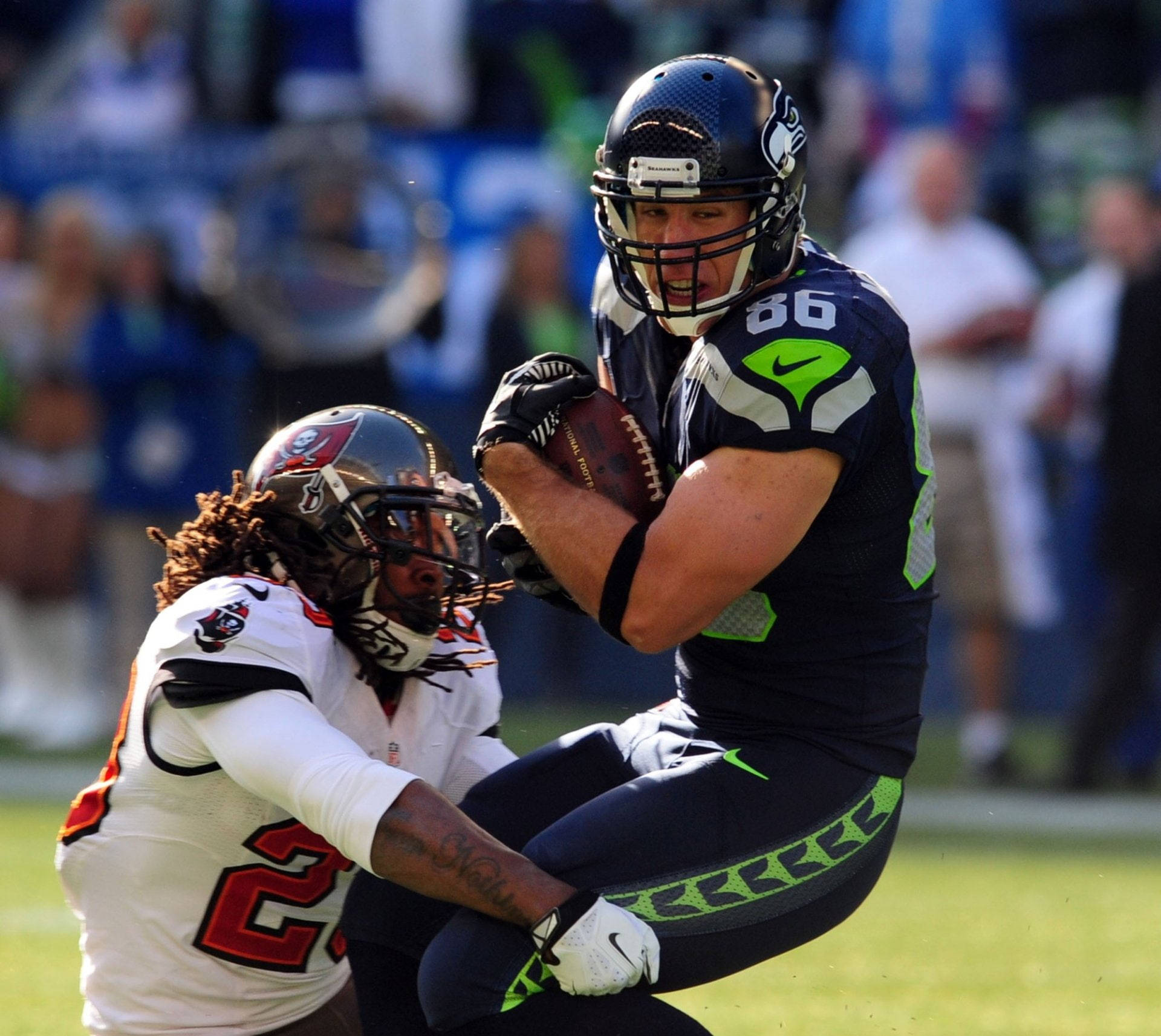 Seattle Seahawks 桌布 壁紙 — 西雅圖海鷹隊的外接手身穿藍綠球衣接住橄欖球，同時被坦帕灣海盜隊身穿白色球衣的防守球員擒抱。